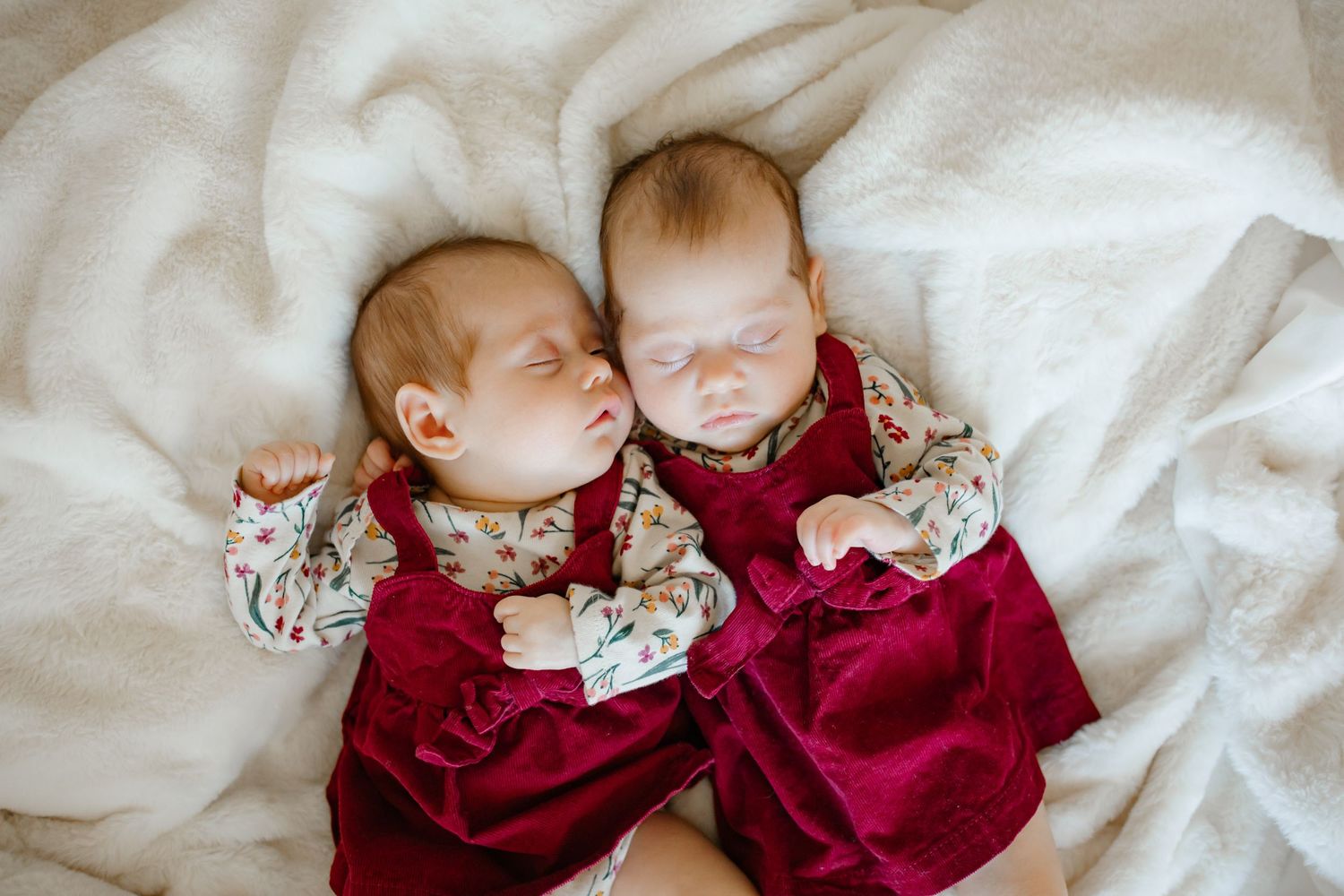 Twin babies in matching red velvet dresses with floral details resting on white fabric.