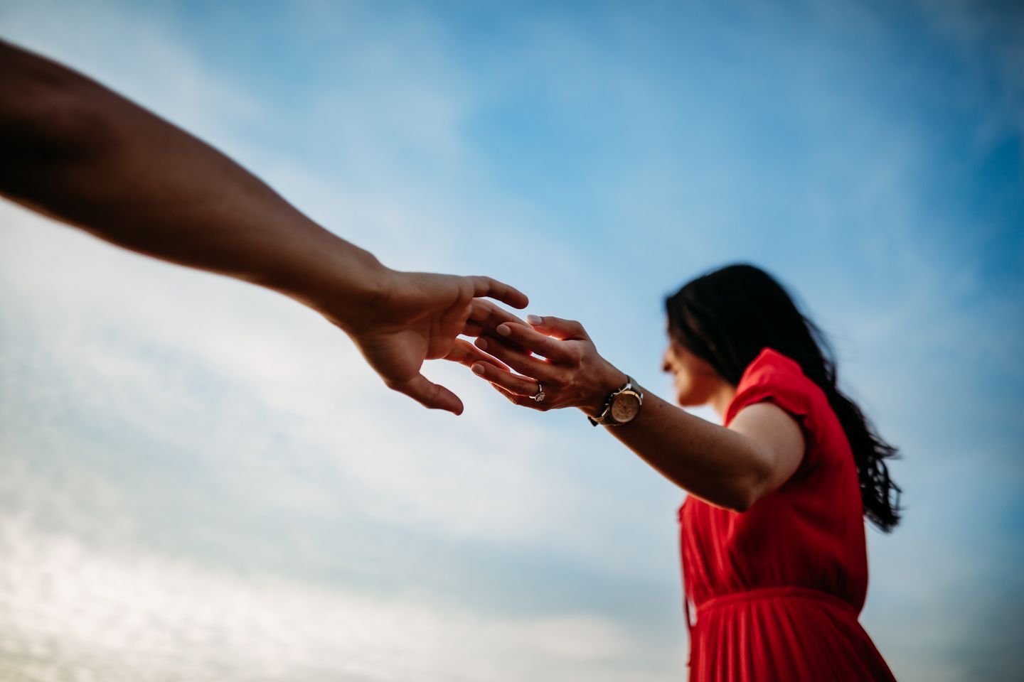 Two hands reaching towards each other against a blue sky background, with a person in a red dress standing below.