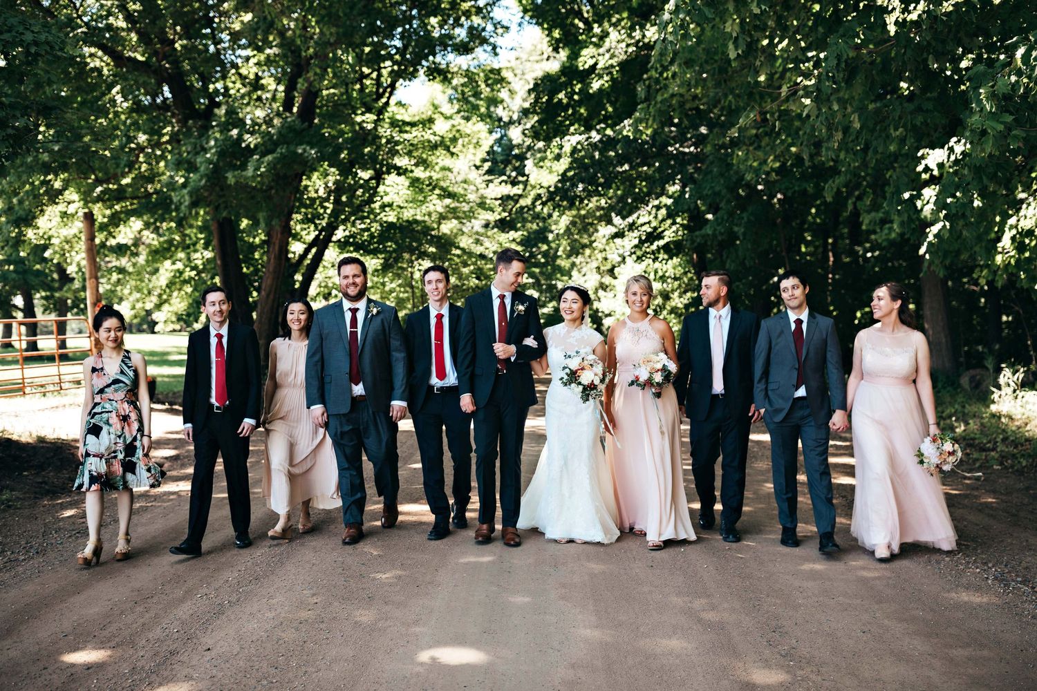 Full wedding party poses on a tree-lined path in coordinated navy suits and pink dresses.