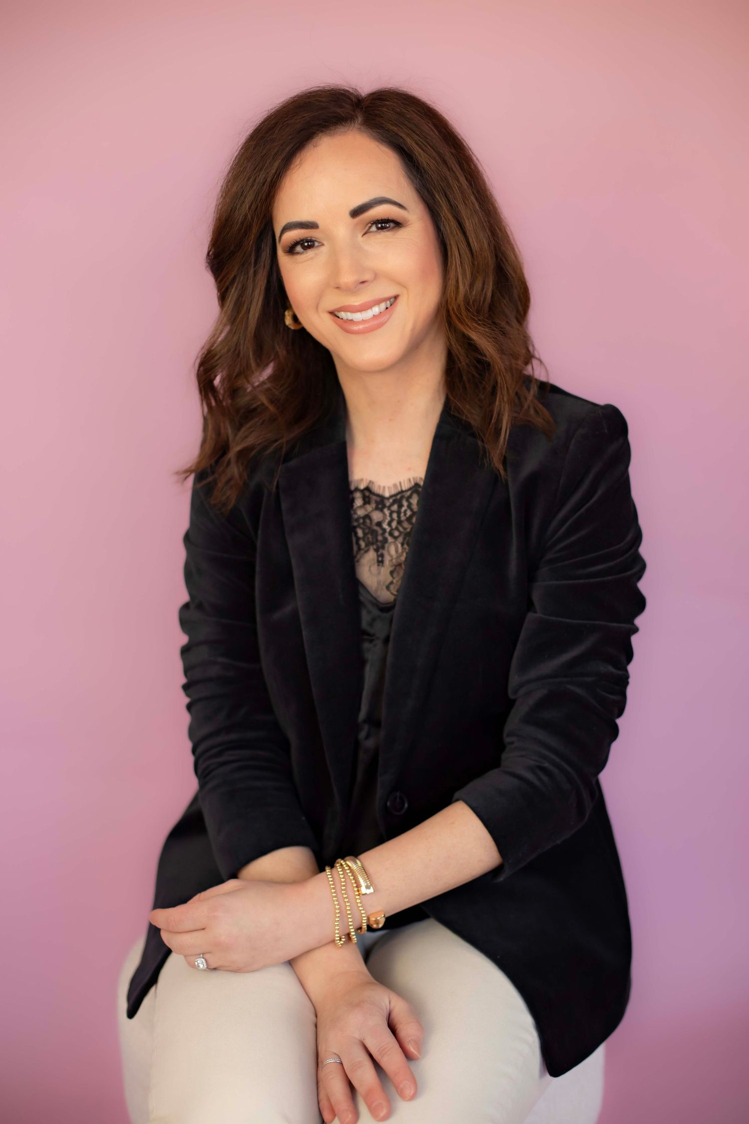 Professional headshot with black velvet blazer and lace top against pink backdrop.