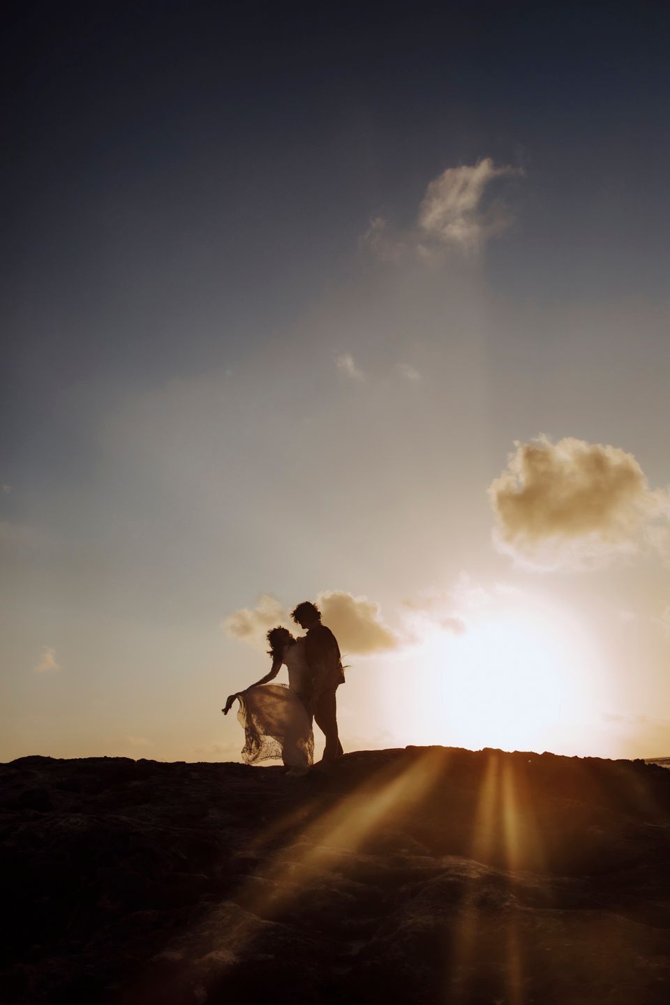 Silhouettes of a couple embrace against a dramatic sunset on a hilltop.