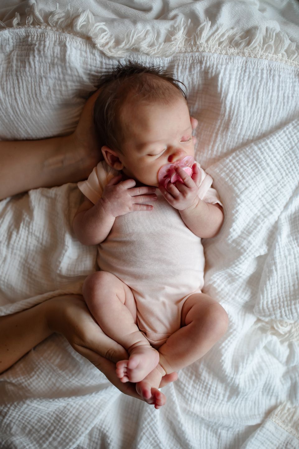 Newborn with pacifier resting peacefully on white textured bedding.