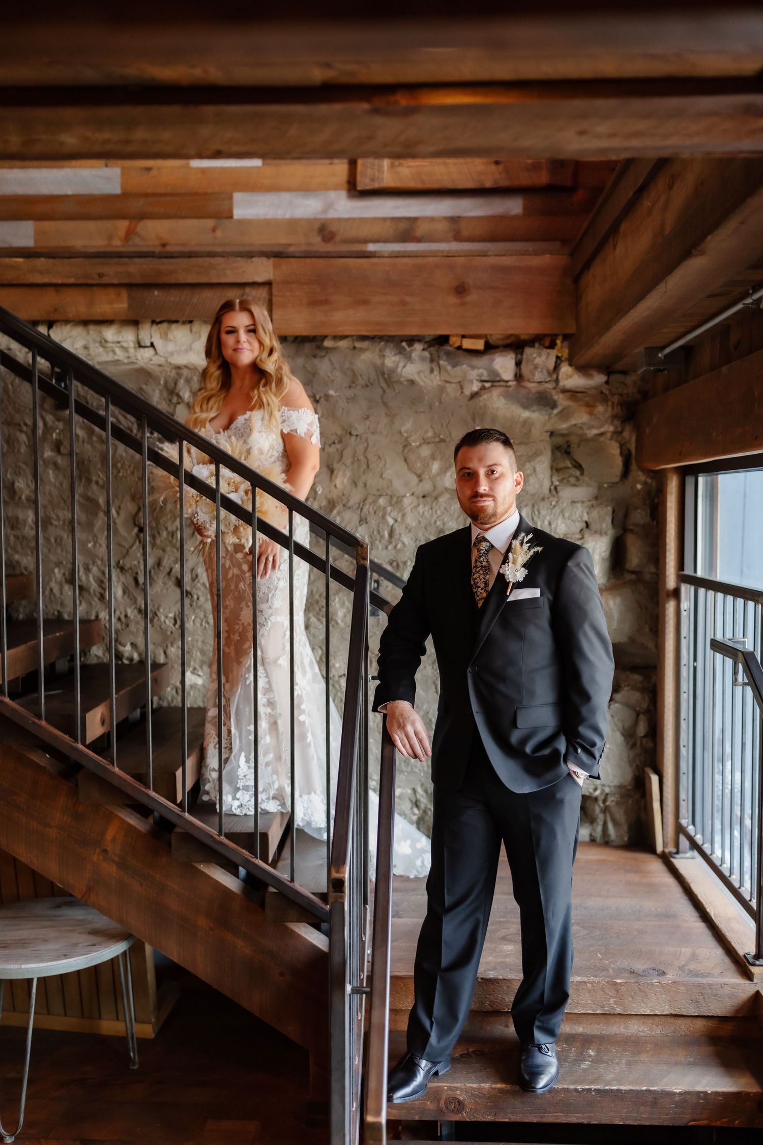 A wedding couple standing on rustic wooden stairs with industrial-style railings and exposed stone walls.
