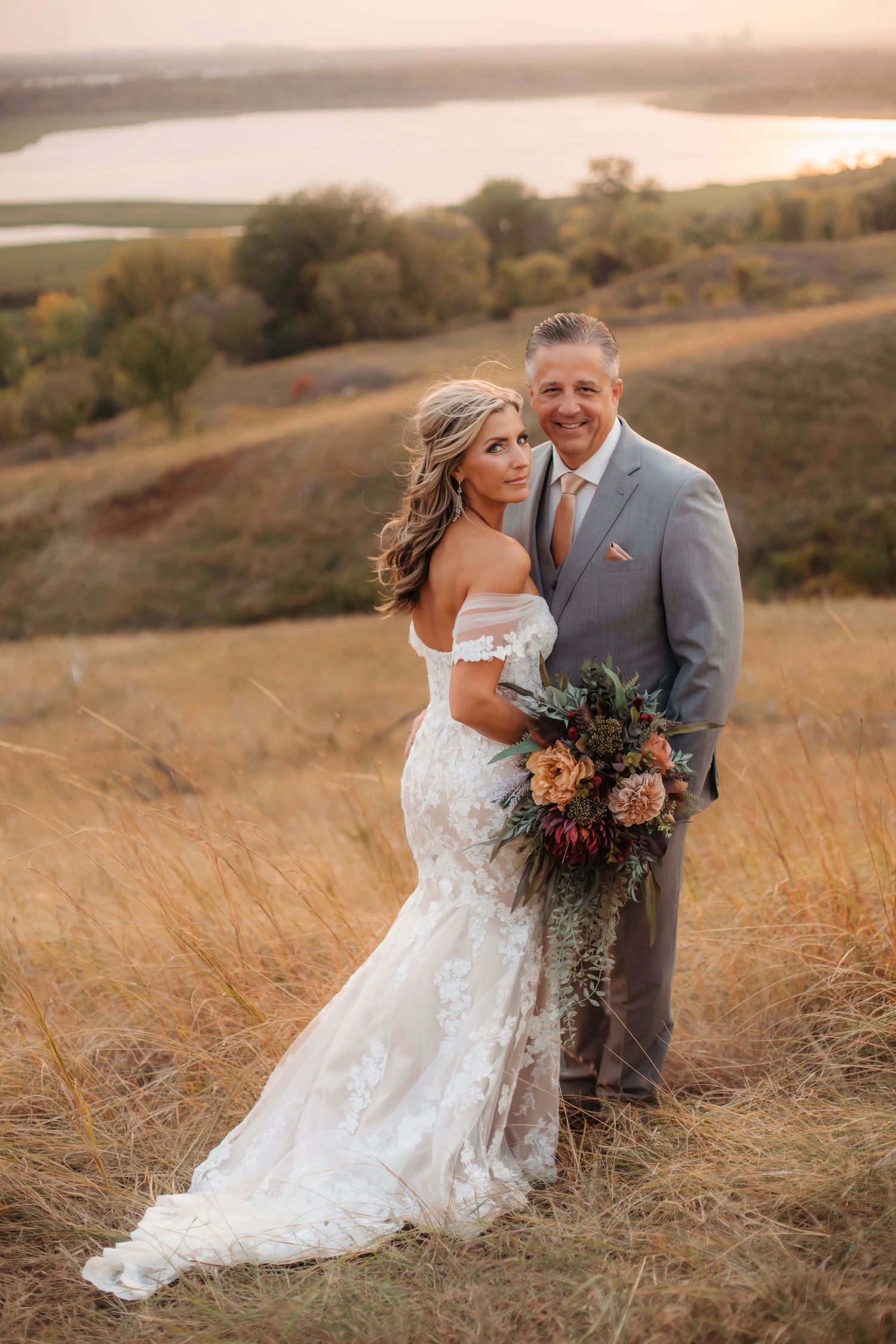 A wedding couple embraces on a hilltop with a scenic lake view during sunset.