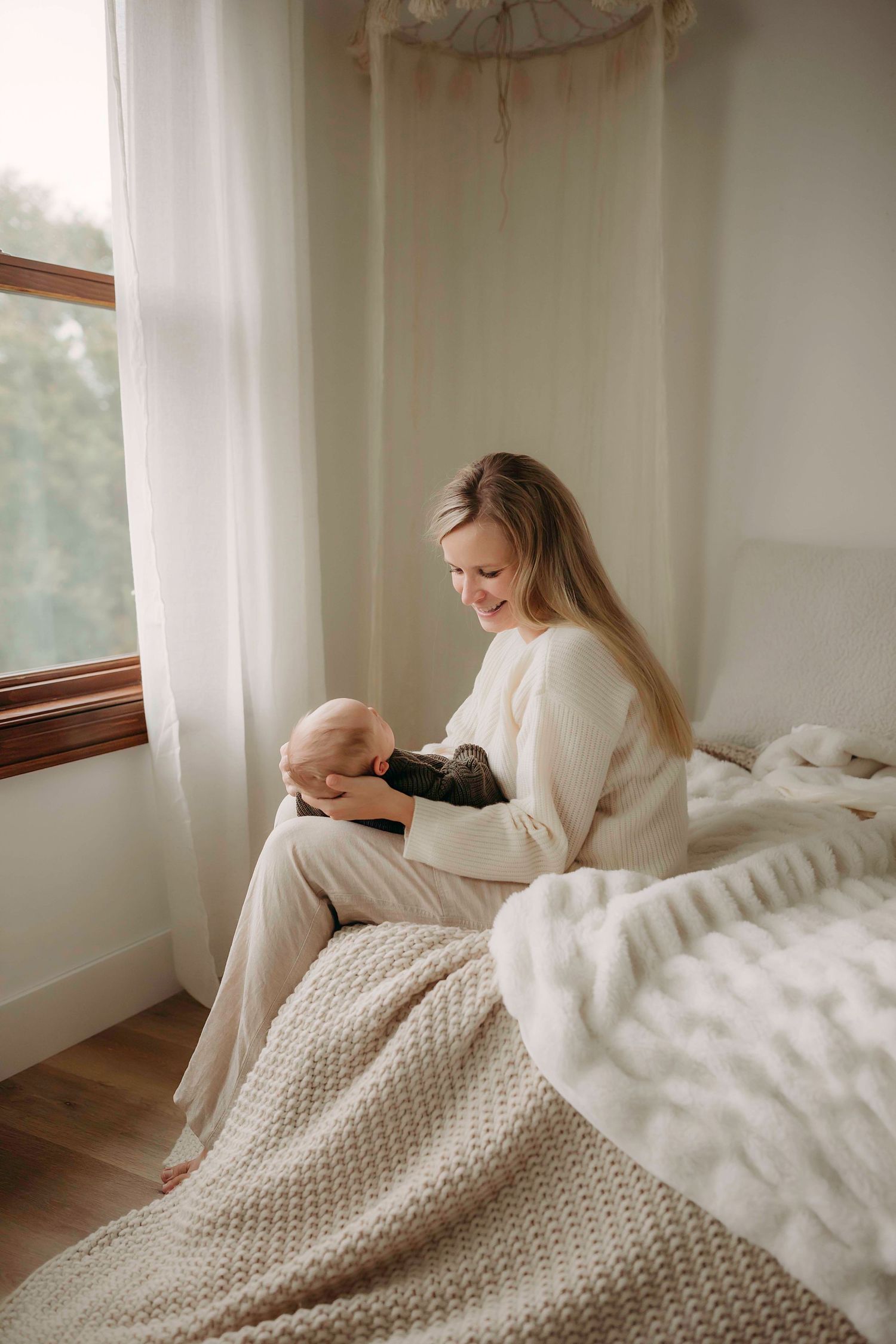 Woman in white sweater sitting by window holding sleeping infant on sunny morning.
