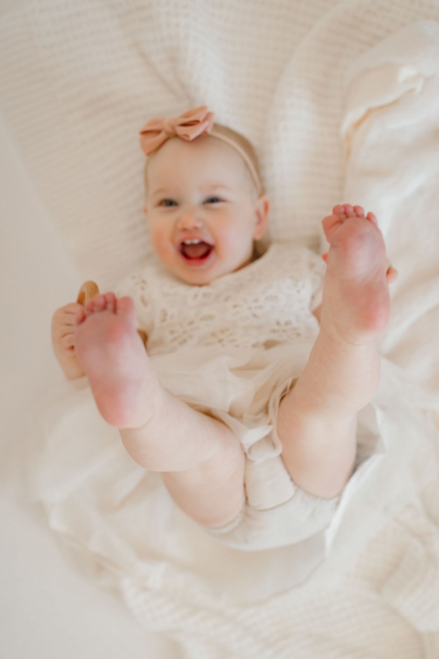 Baby in white dress with bow smiling and showing bare feet on white blanket.