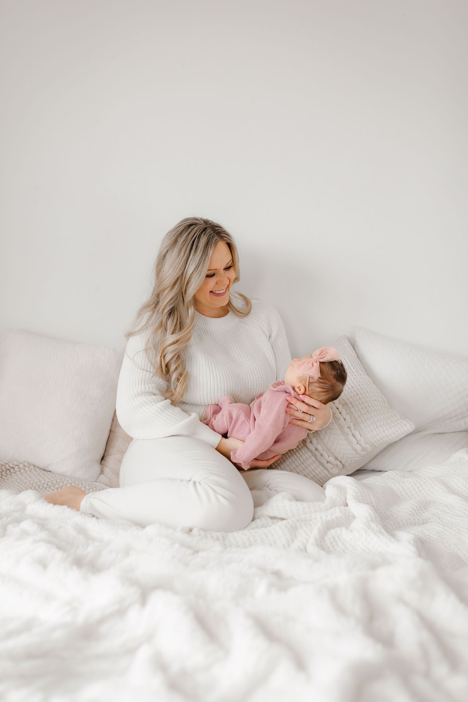 Person in white sweater holding sleeping baby on cozy white bed.