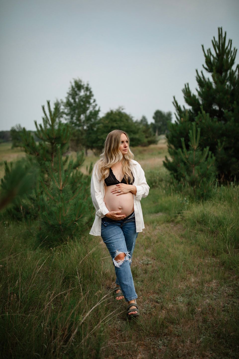 A casual outdoor maternity portrait showing an expectant mother wearing ripped jeans and a white shirt among pine trees.