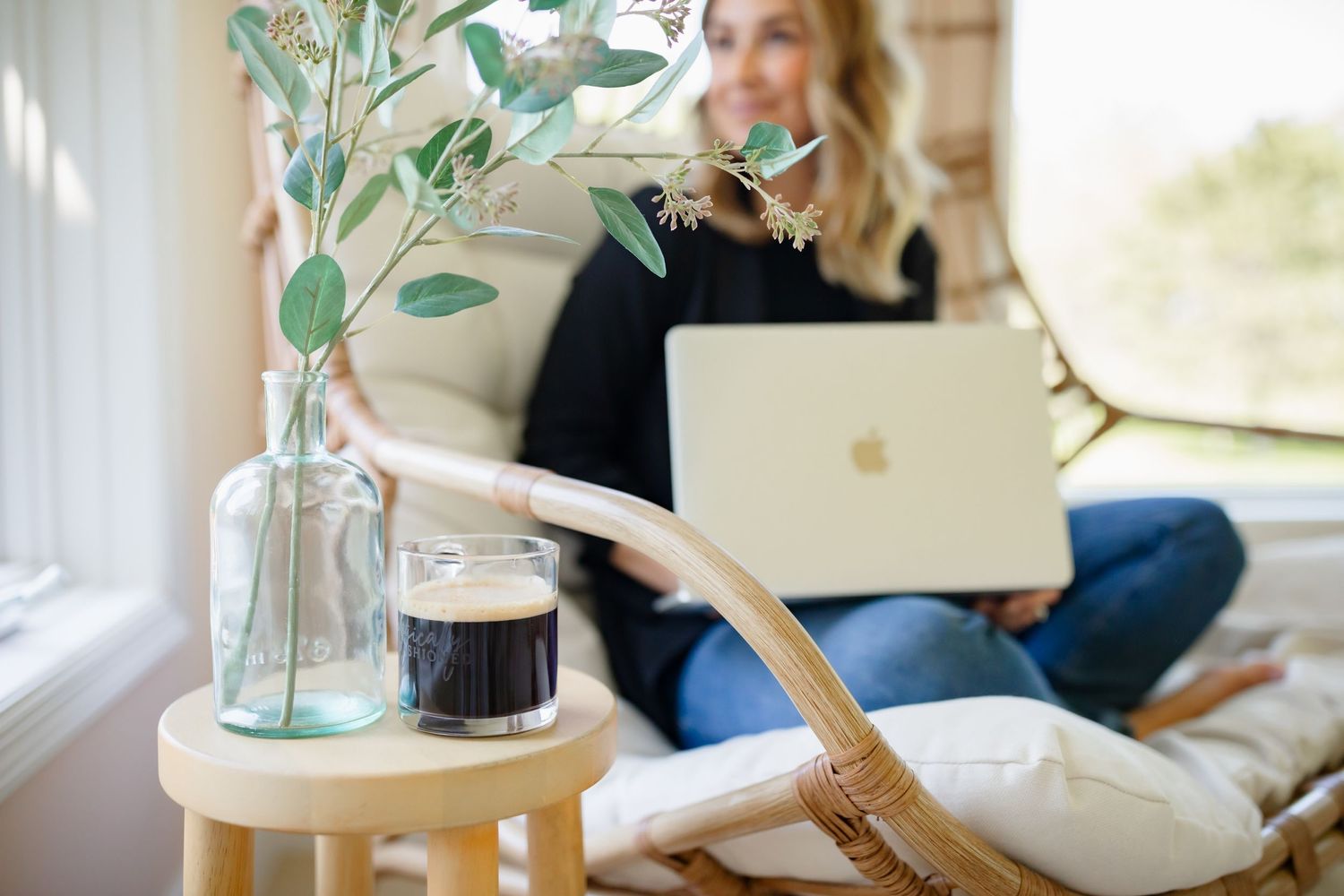 Close-up of MacBook laptop, glass vase with greenery, and coffee on wooden side table near rattan chair.