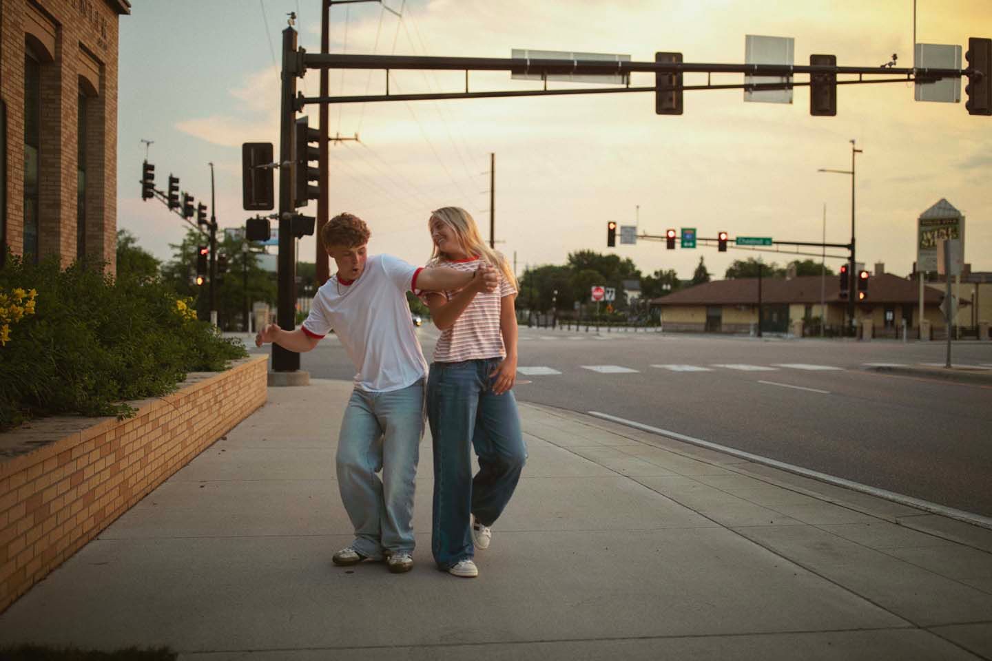 Two friends walk arm in arm along a sidewalk at a quiet intersection during golden hour in a small town.