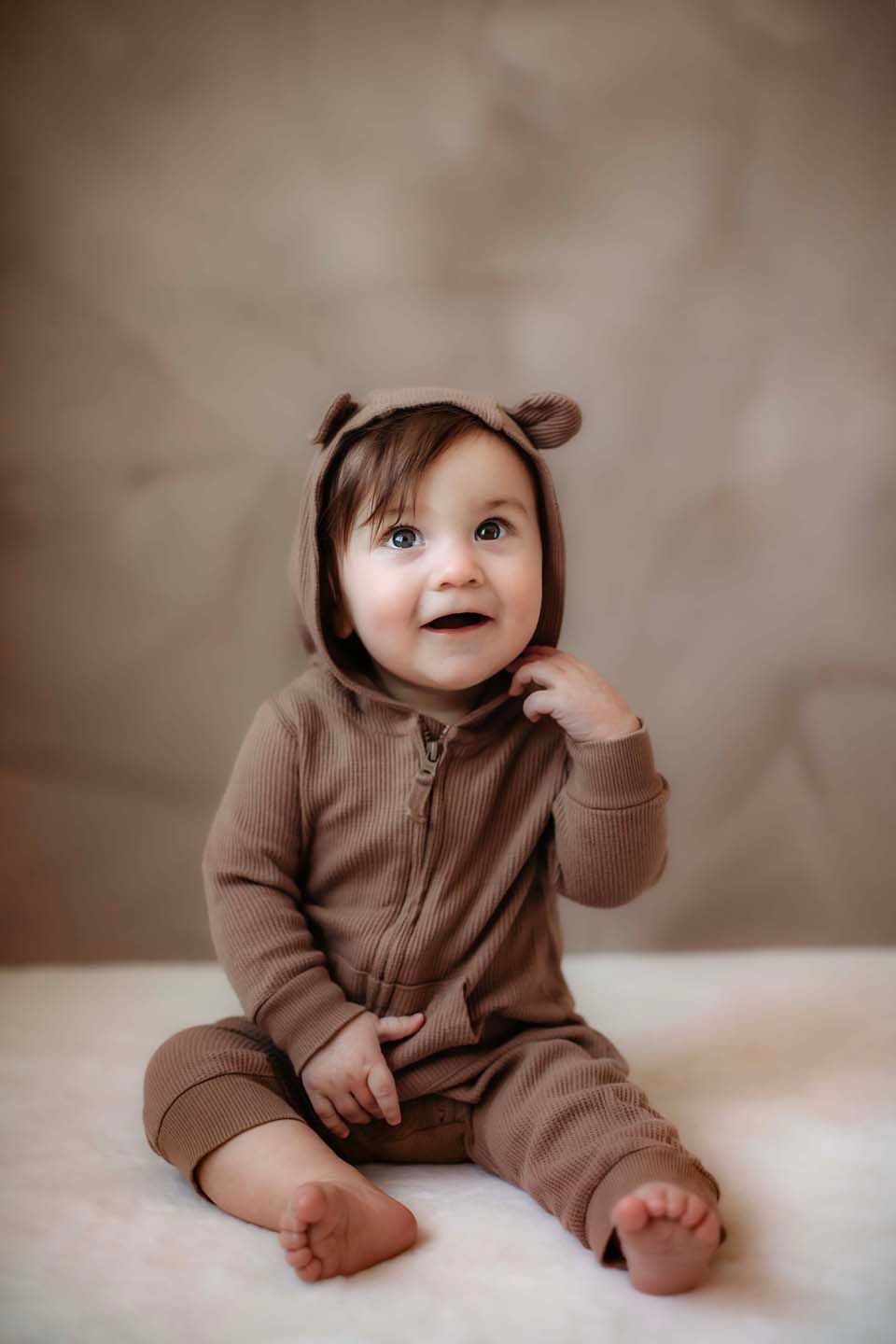 A series of photos showing a baby in a brown hooded onesie sitting on a white surface.