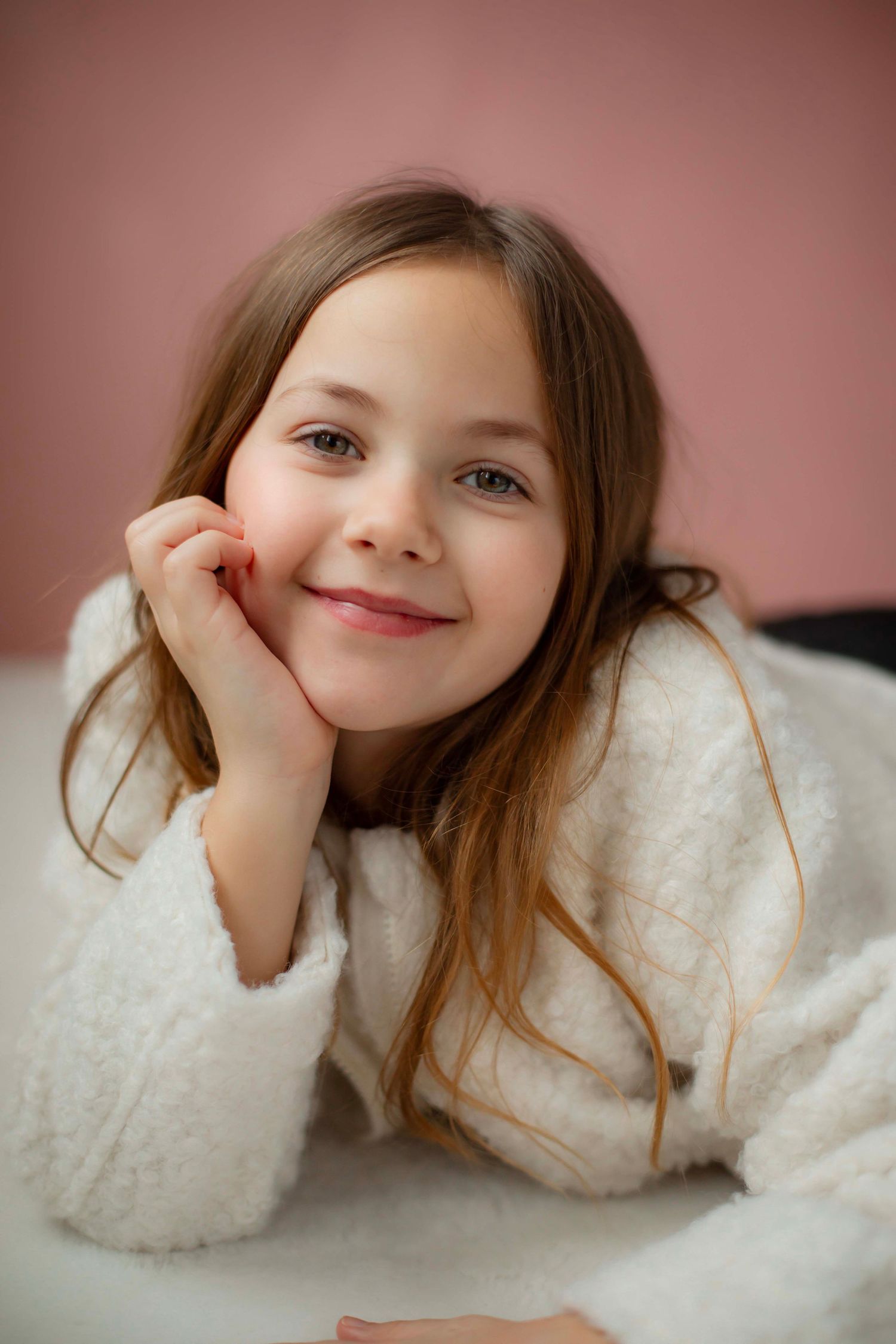 A child wearing a cozy white sweater poses playfully while lying down against a pink background.
