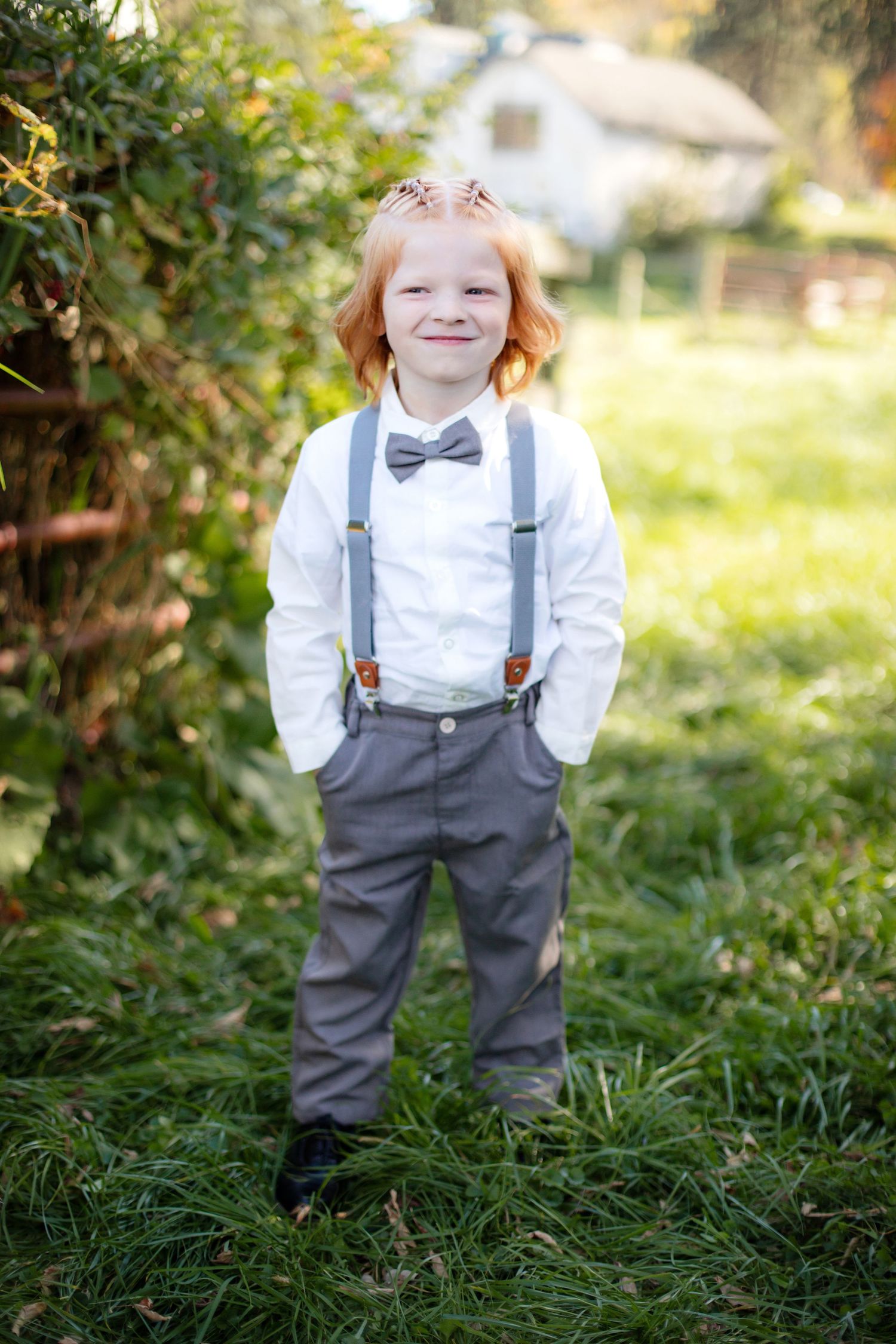 A young attendant in suspenders, bowtie and formal wear smiles while standing in a garden setting.