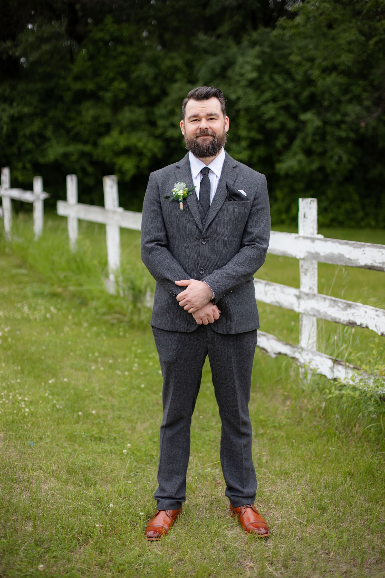 Groom in a charcoal suit stands by a white fence on a grassy lawn.