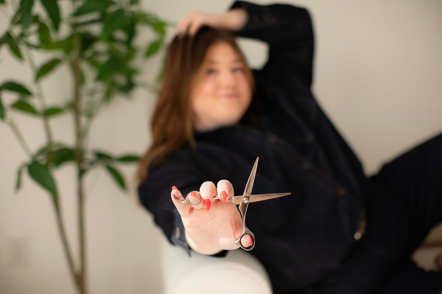 Woman in black outfit holding scissors toward camera in a bright salon setting with green plants in background.