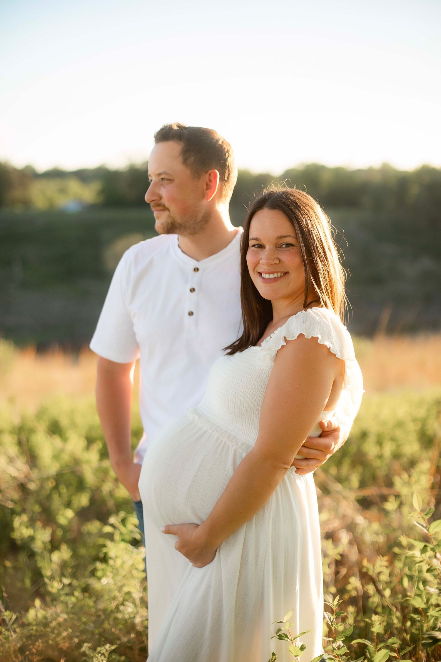 Smiling pregnant woman in white dress with partner standing in sunny field during golden hour maternity photo shoot.