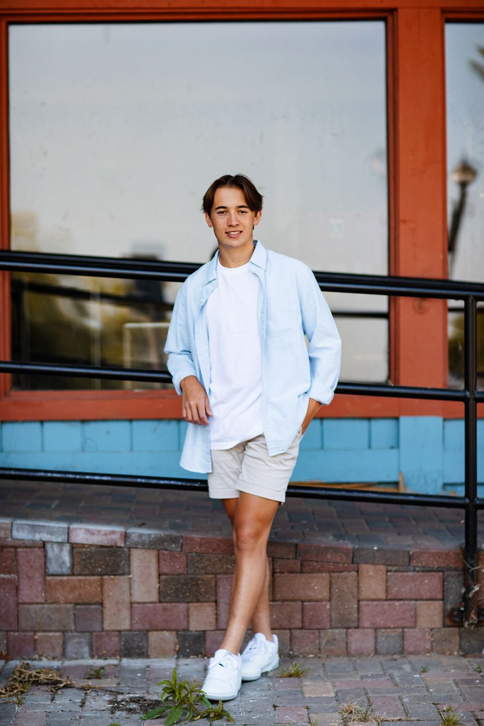 Casual summer fashion with light blue button-up shirt, white t-shirt, beige shorts and white sneakers against brick wall.