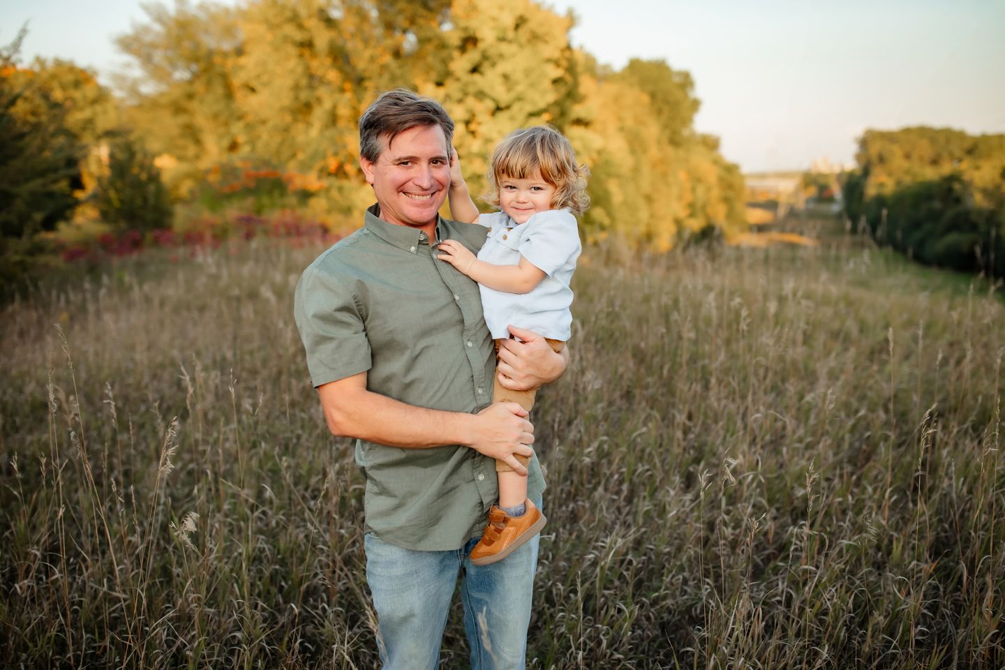 Loving parent holds their young child while standing in a golden sunlit meadow at sunset.