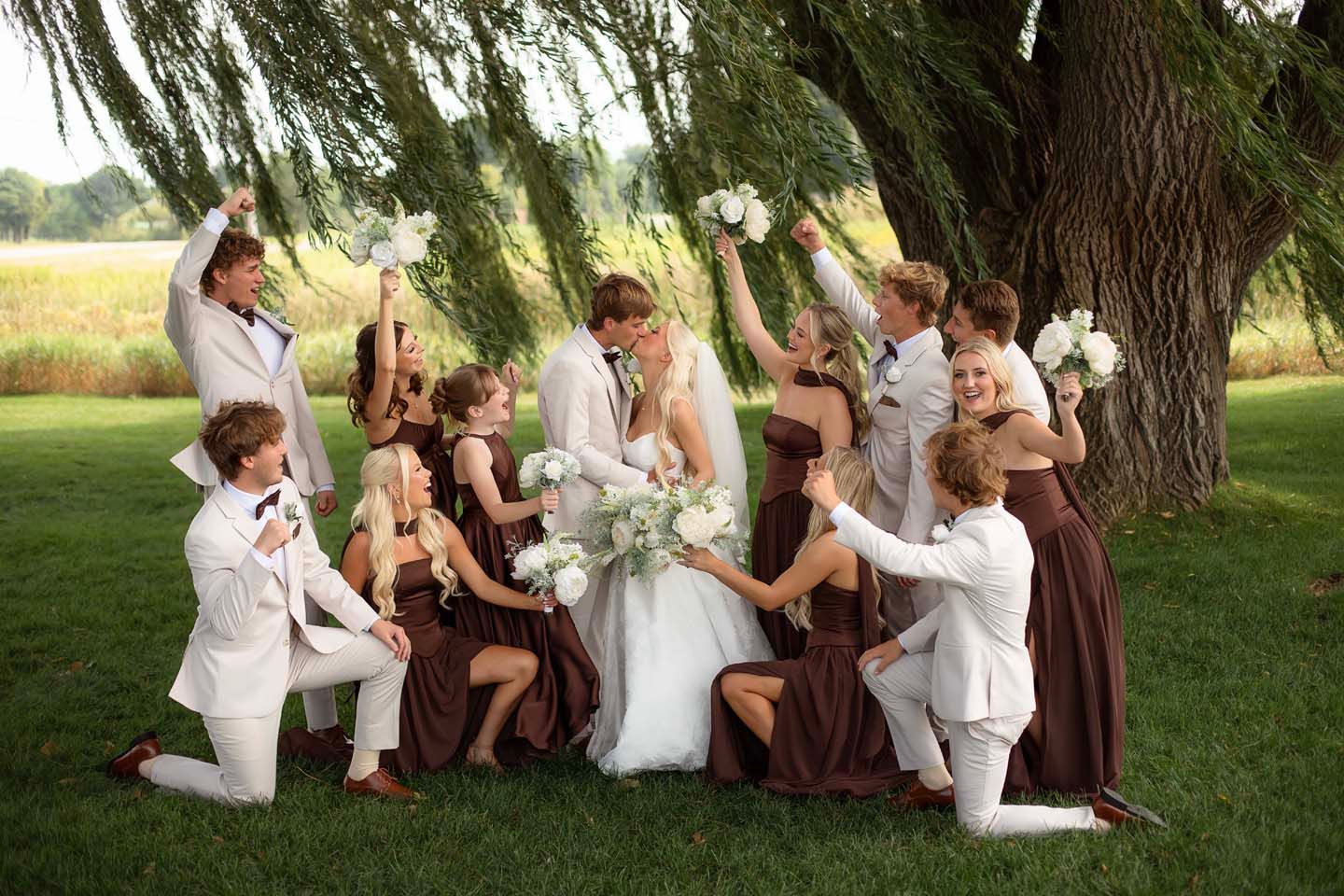 Wedding party celebrates bride and groom under willow tree, bridesmaids in brown dresses and groomsmen in white suits.