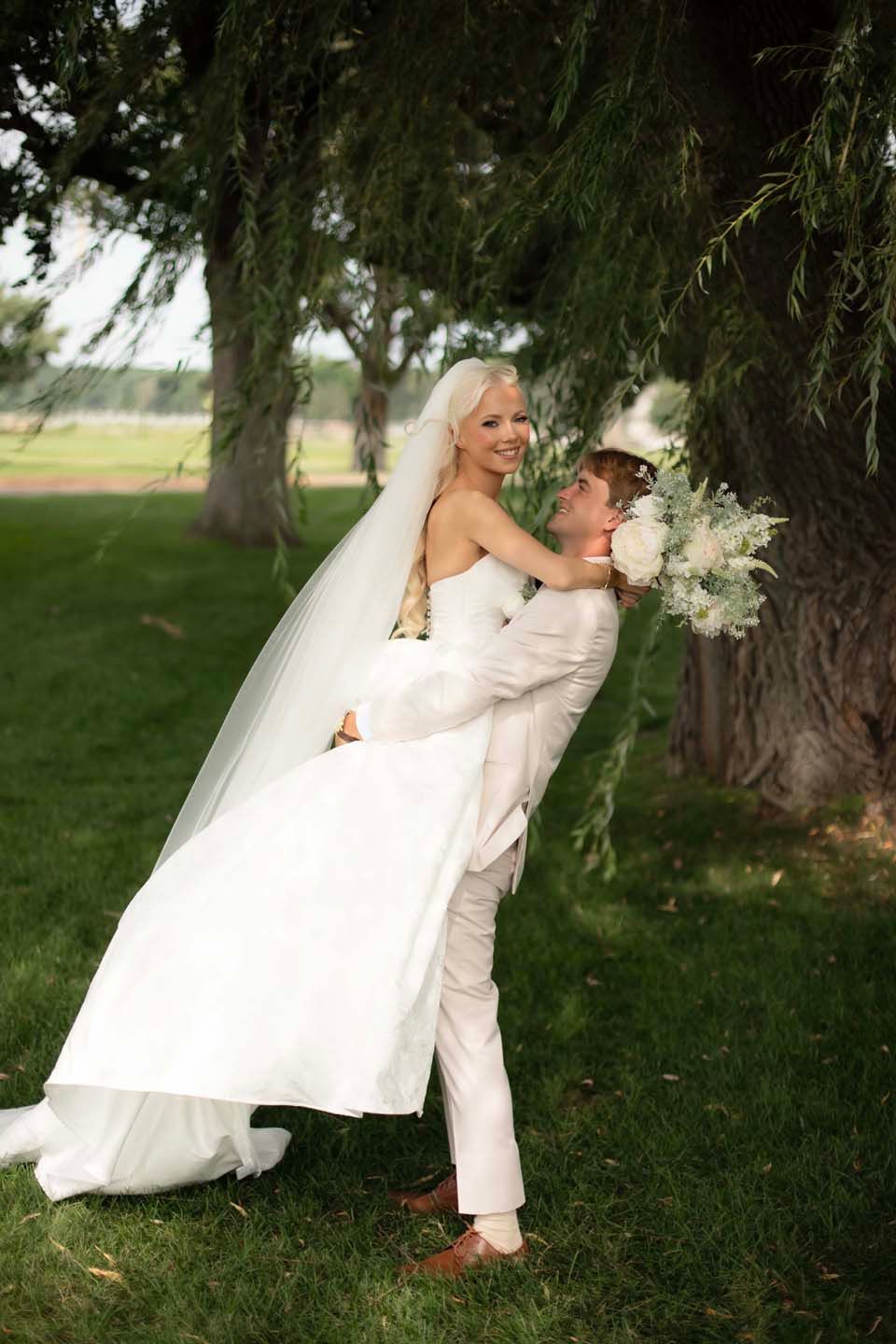 Groom lifts smiling bride in white gown and veil outdoors under a large green tree.