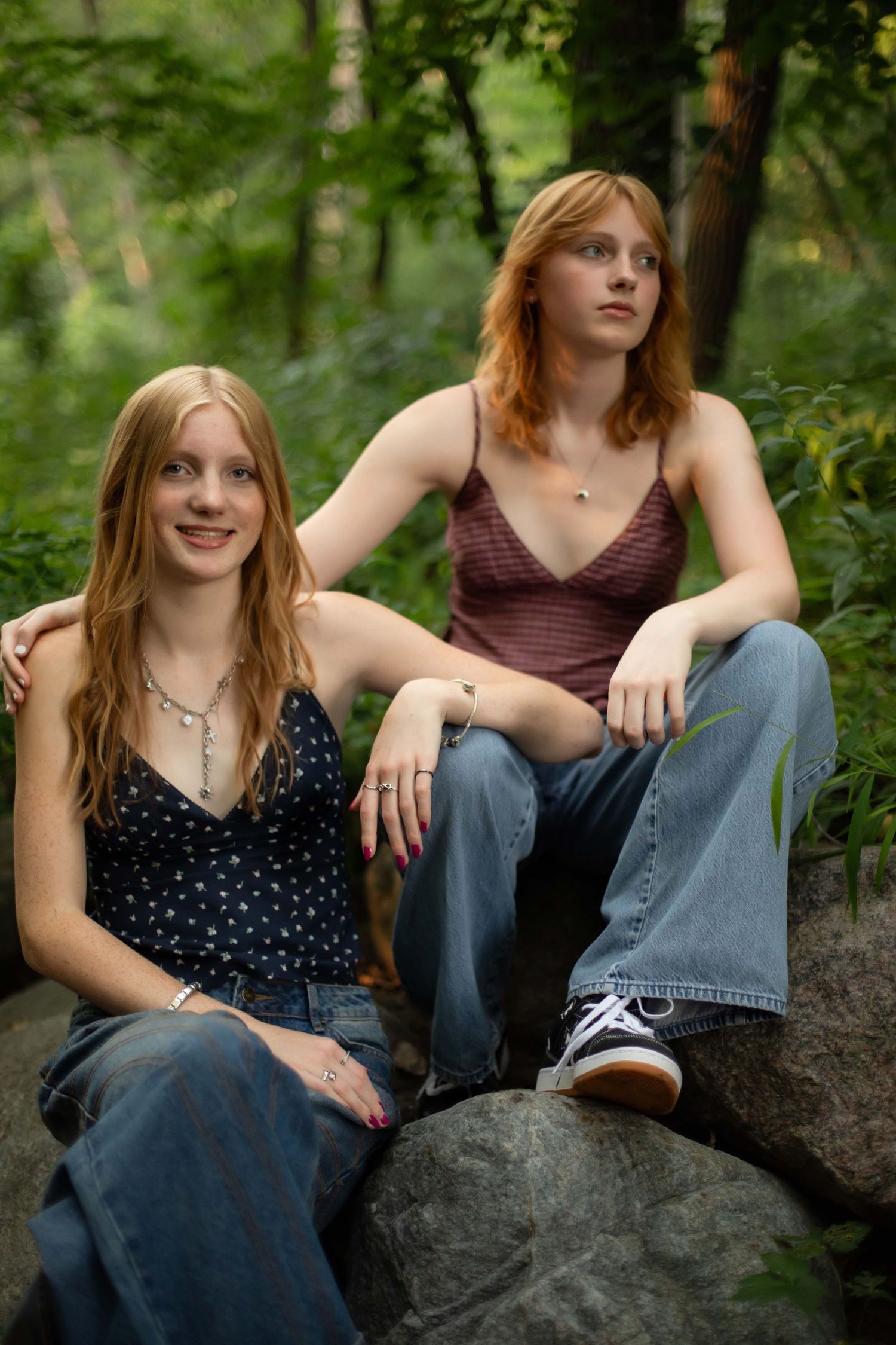 Two teenage girls with red hair sitting on rocks in a lush green forest, wearing jeans and casual tops.