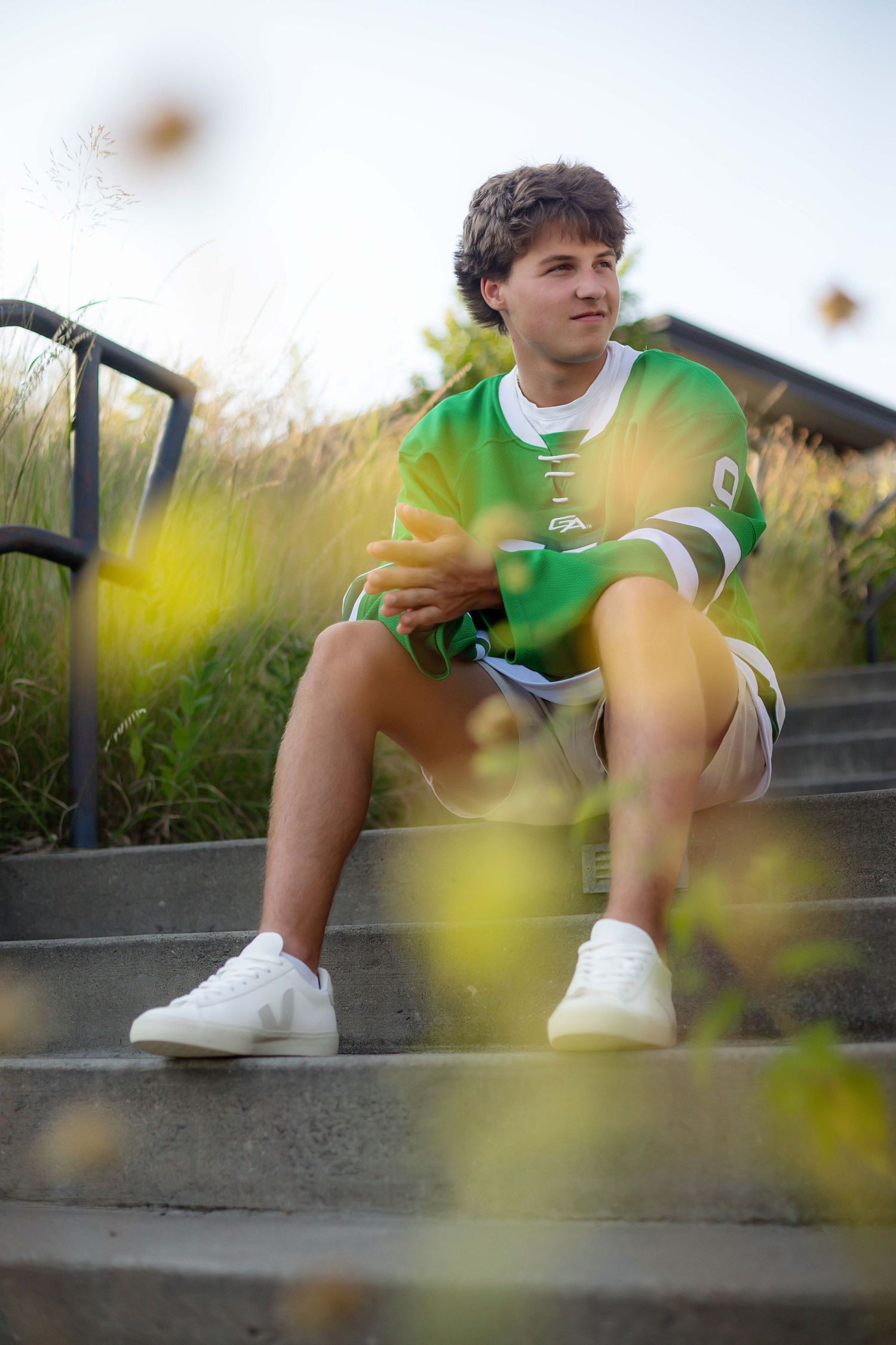 Young man in green sports jersey and white sneakers sitting on outdoor steps surrounded by tall grass.
