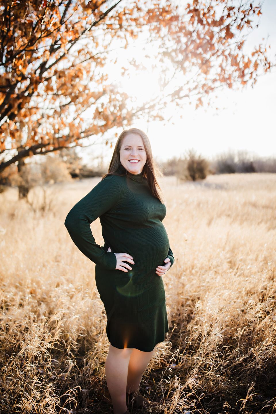 Beautiful maternity photo in wheat field during golden hour with autumn trees in background, wearing dark green dress.