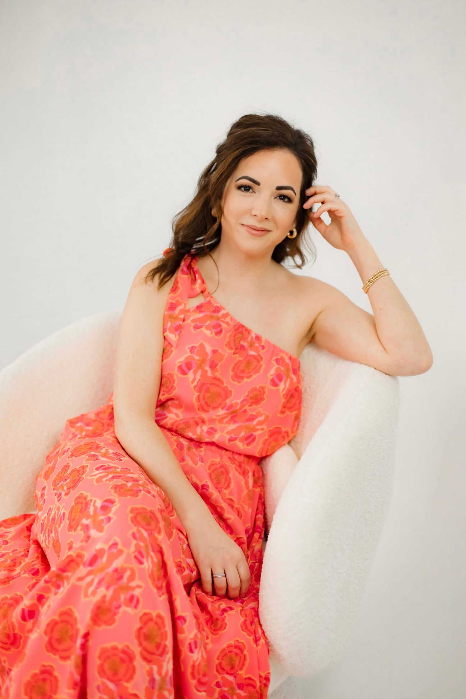 Woman in coral floral one-shoulder dress poses elegantly on white chair.