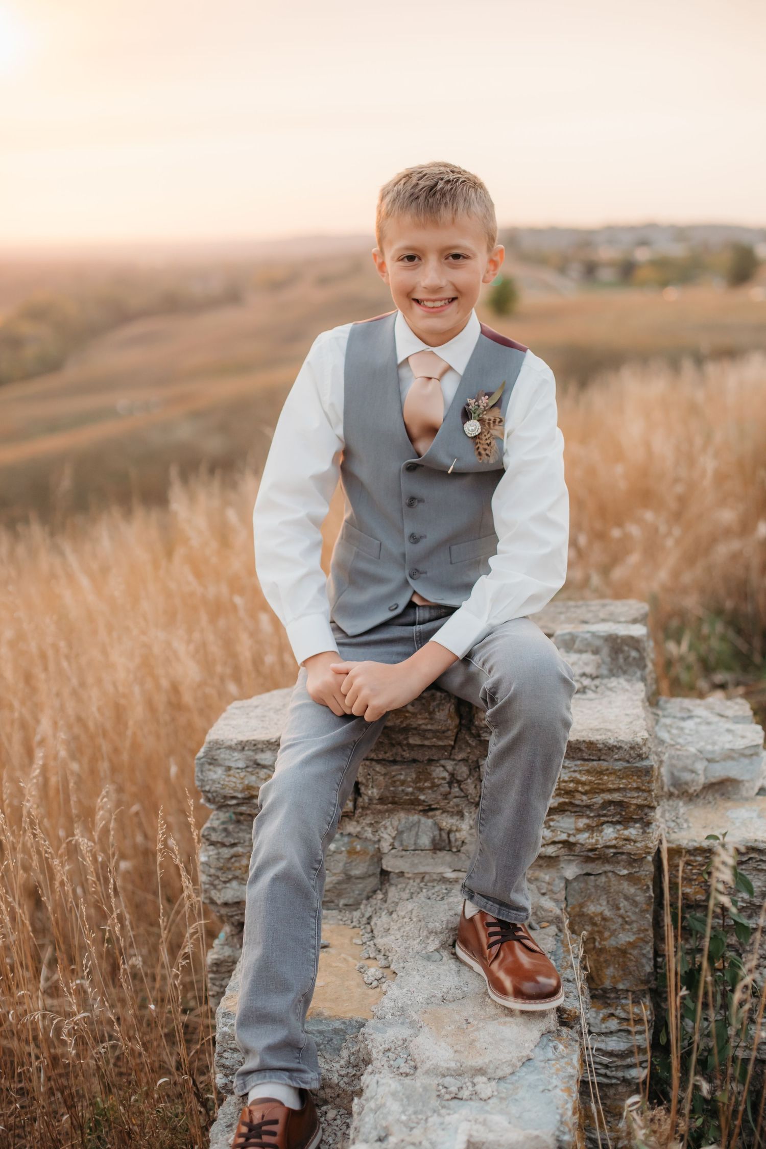 Boy in grey vest and tie sits on stone wall during sunset portrait session.