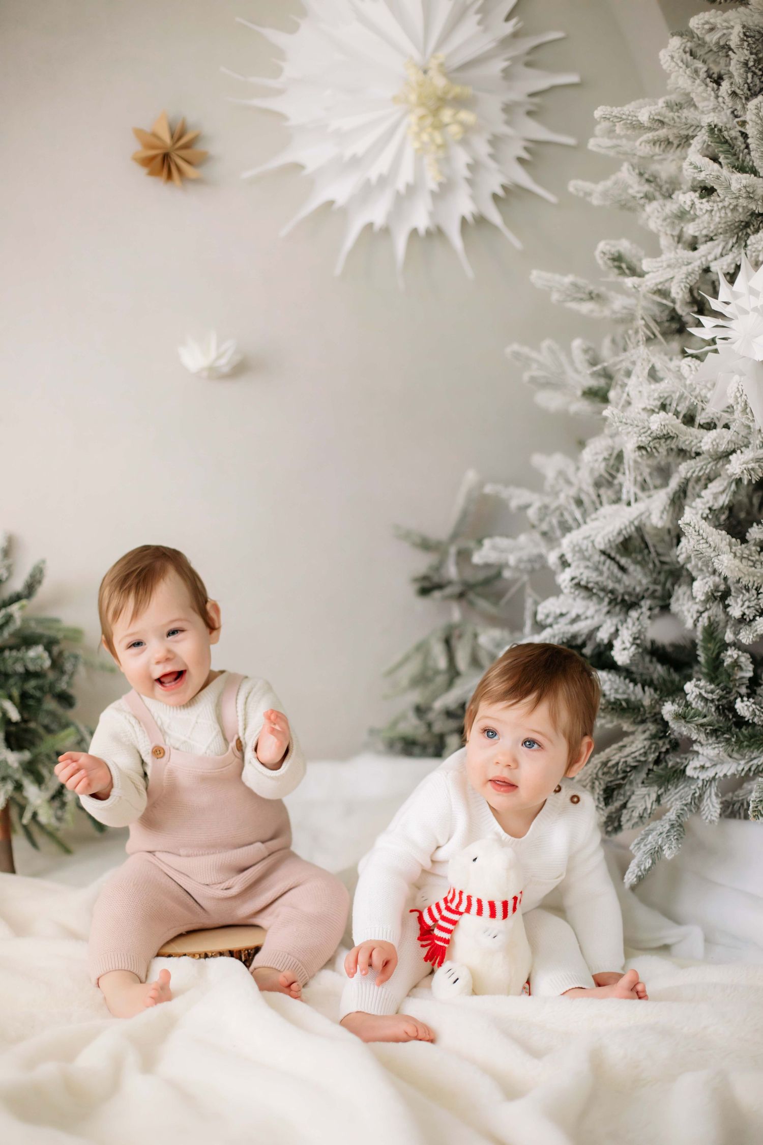 Two babies sit on white blanket near flocked Christmas tree with festive decorations.