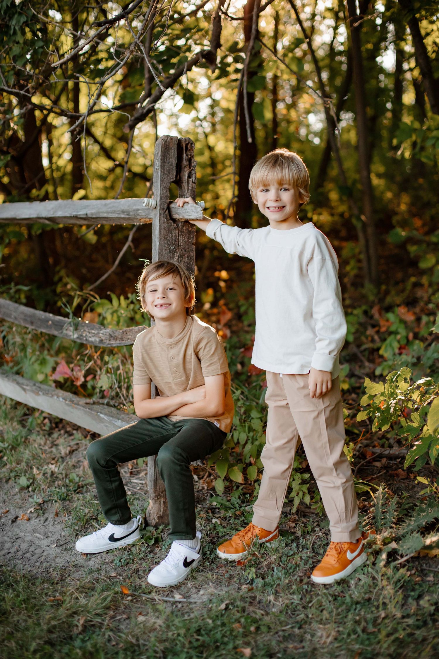 Two young children pose by wooden fence in autumn forest setting.