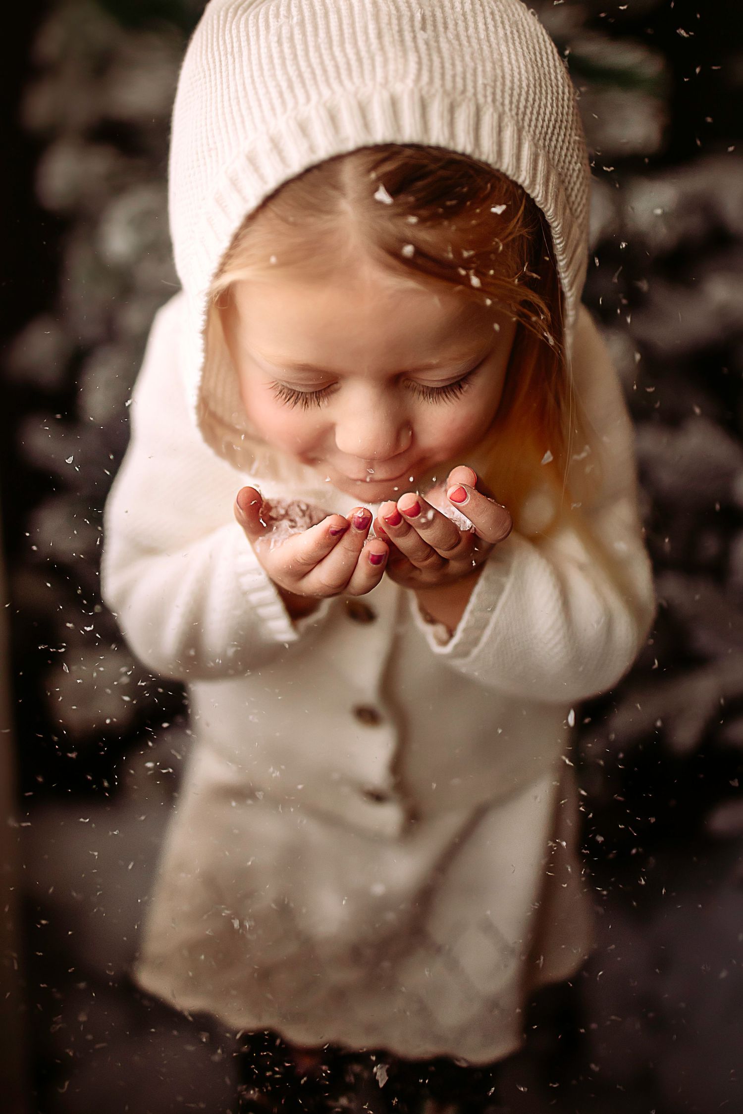 Winter portrait of child in white coat and beanie blowing magical snow in outdoor setting.