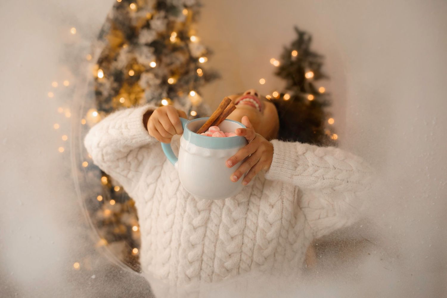 Hands holding a white mug against a backdrop of glowing Christmas trees.