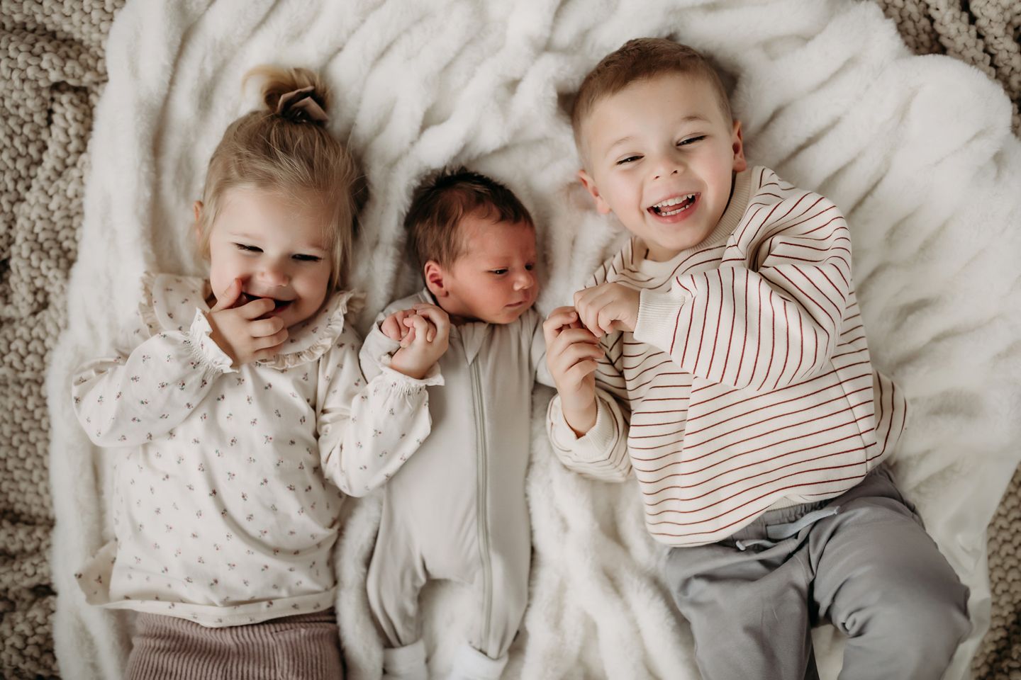 Three siblings lying together on white blanket with joyful expressions.