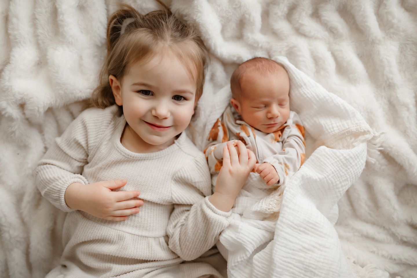 Siblings cuddling on cozy white bedding while wearing matching light colored sweaters.