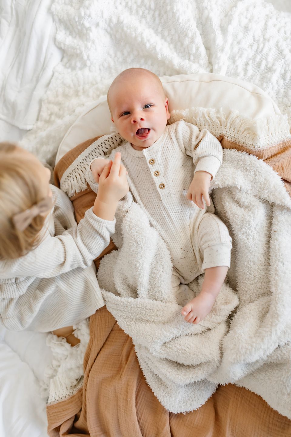 Baby lying on soft white bedding wearing cozy cream-colored outfit while being comforted.