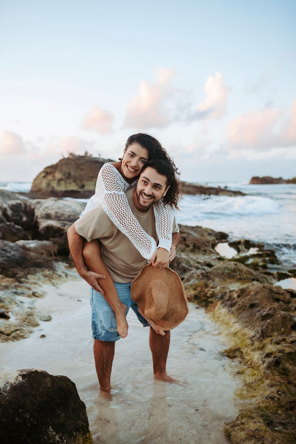 Couple sharing joyful piggyback ride by ocean rocks during sunset.