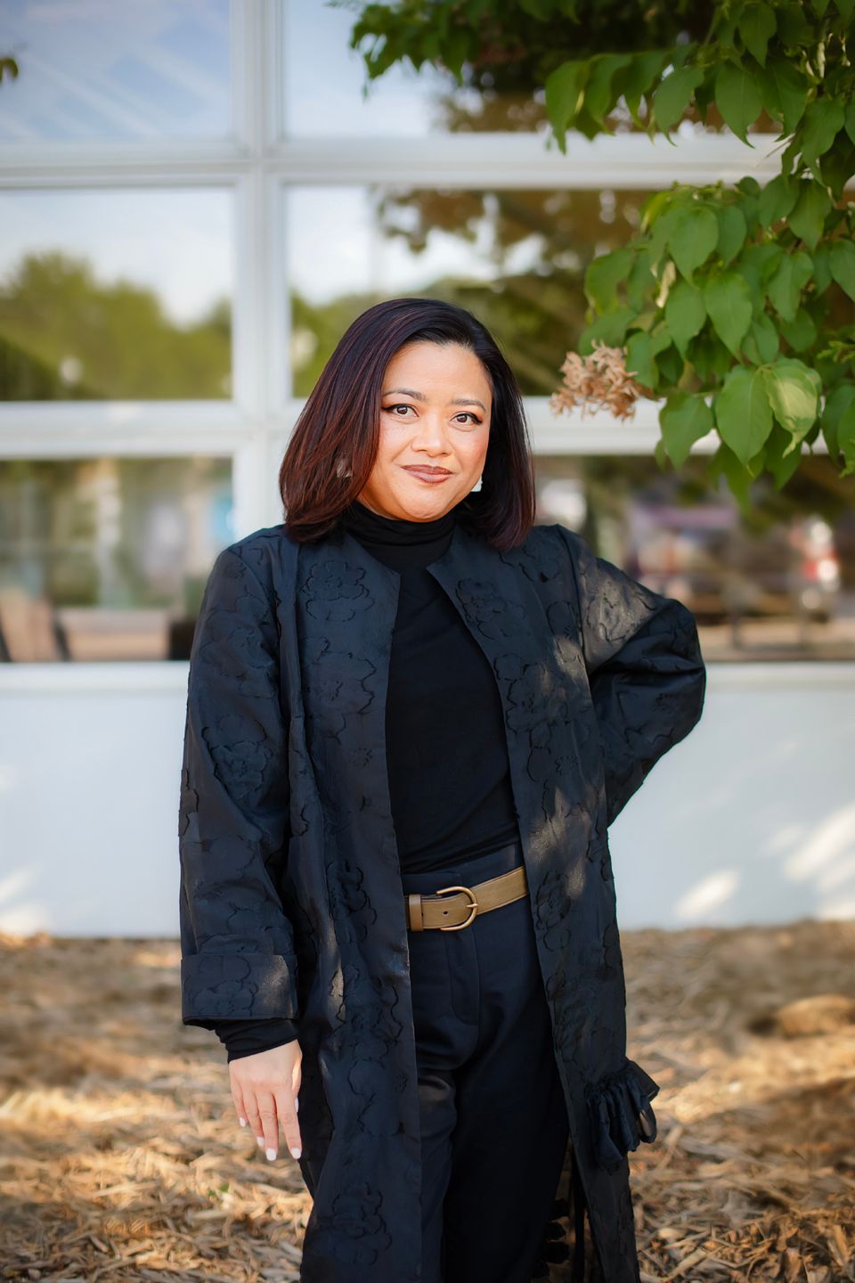 A person in a black blazer and dark outfit poses outdoors against a natural backdrop with trees and sunlight.