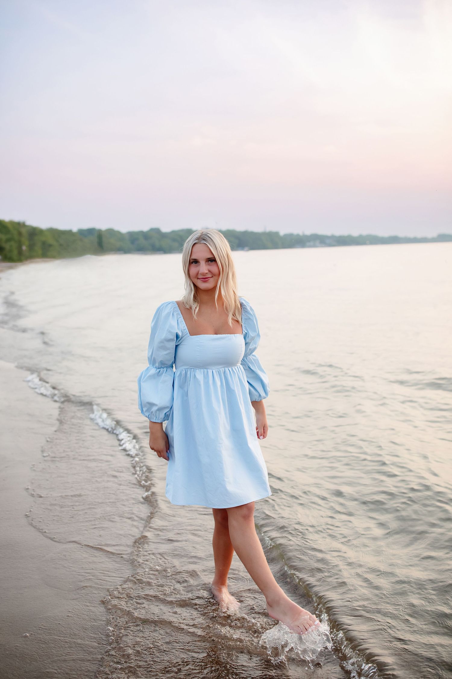 Senior high school in light blue dress walking barefoot along a sandy shoreline at sunset.
