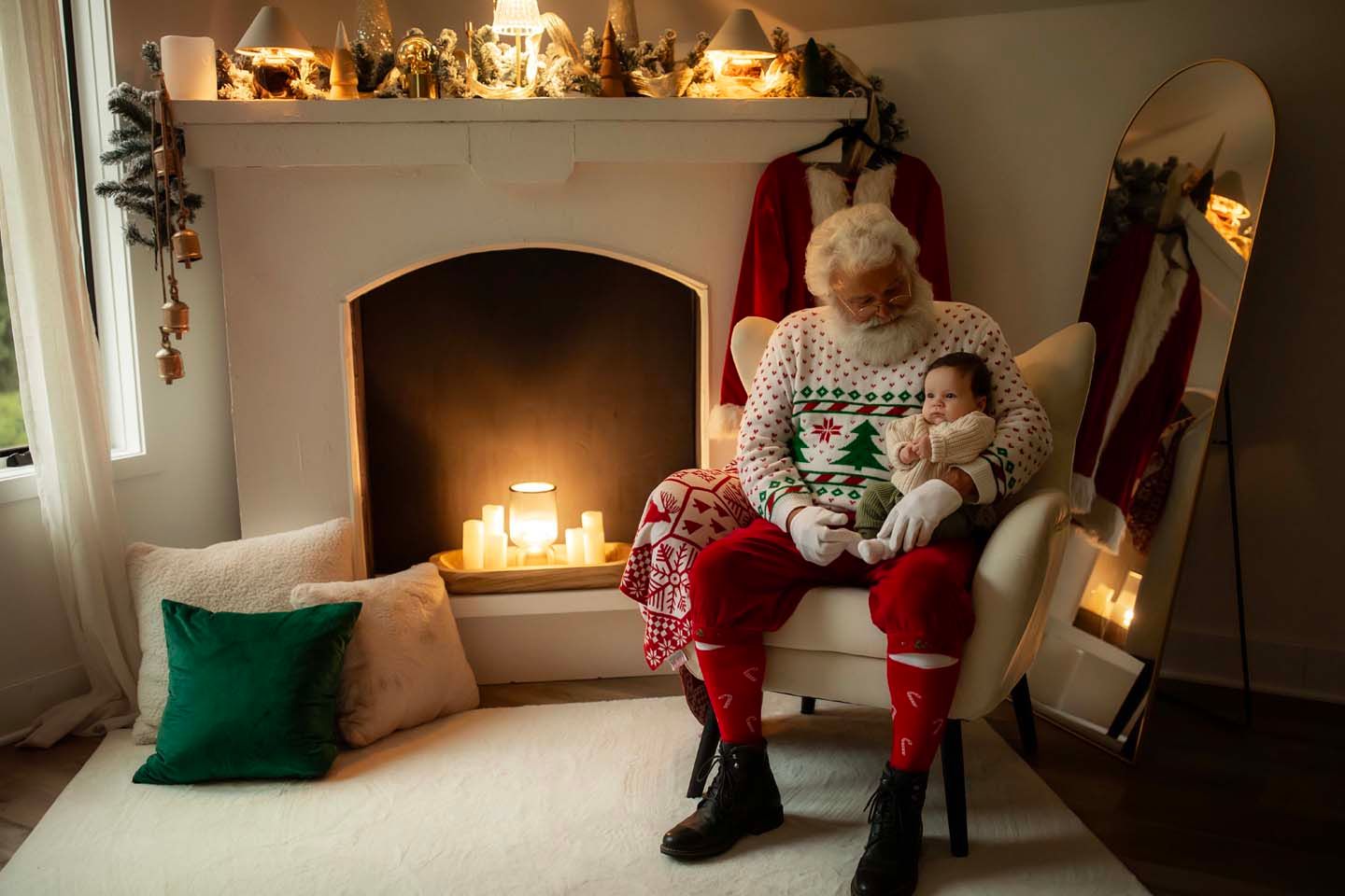 Santa Claus sits in armchair holding sleeping baby near decorated fireplace with candles in cozy Christmas living room.