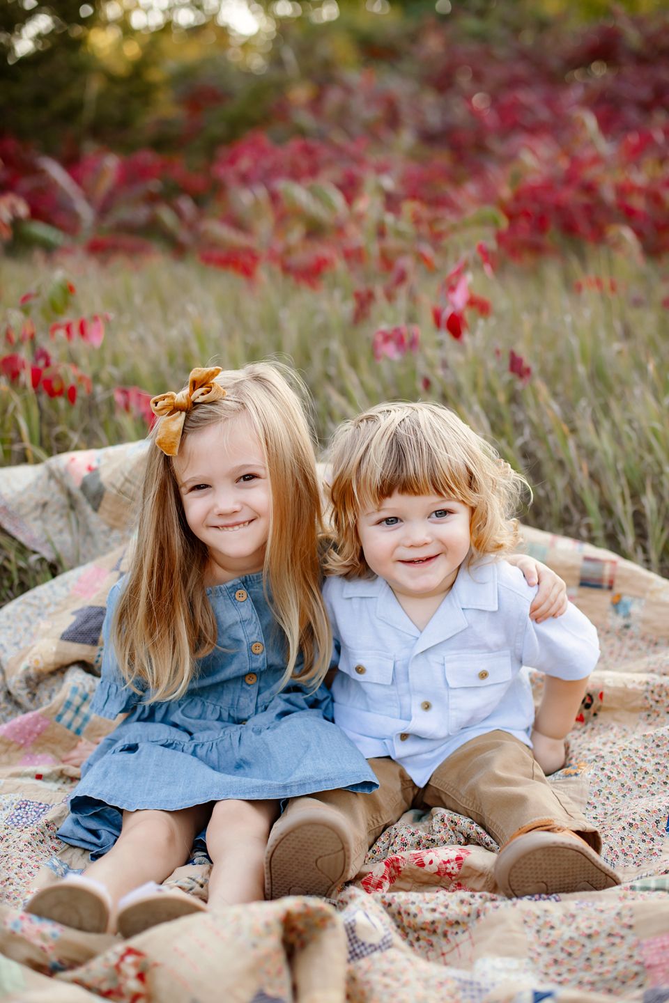 Two young children sitting on a blanket outdoors with autumn red foliage in the background.