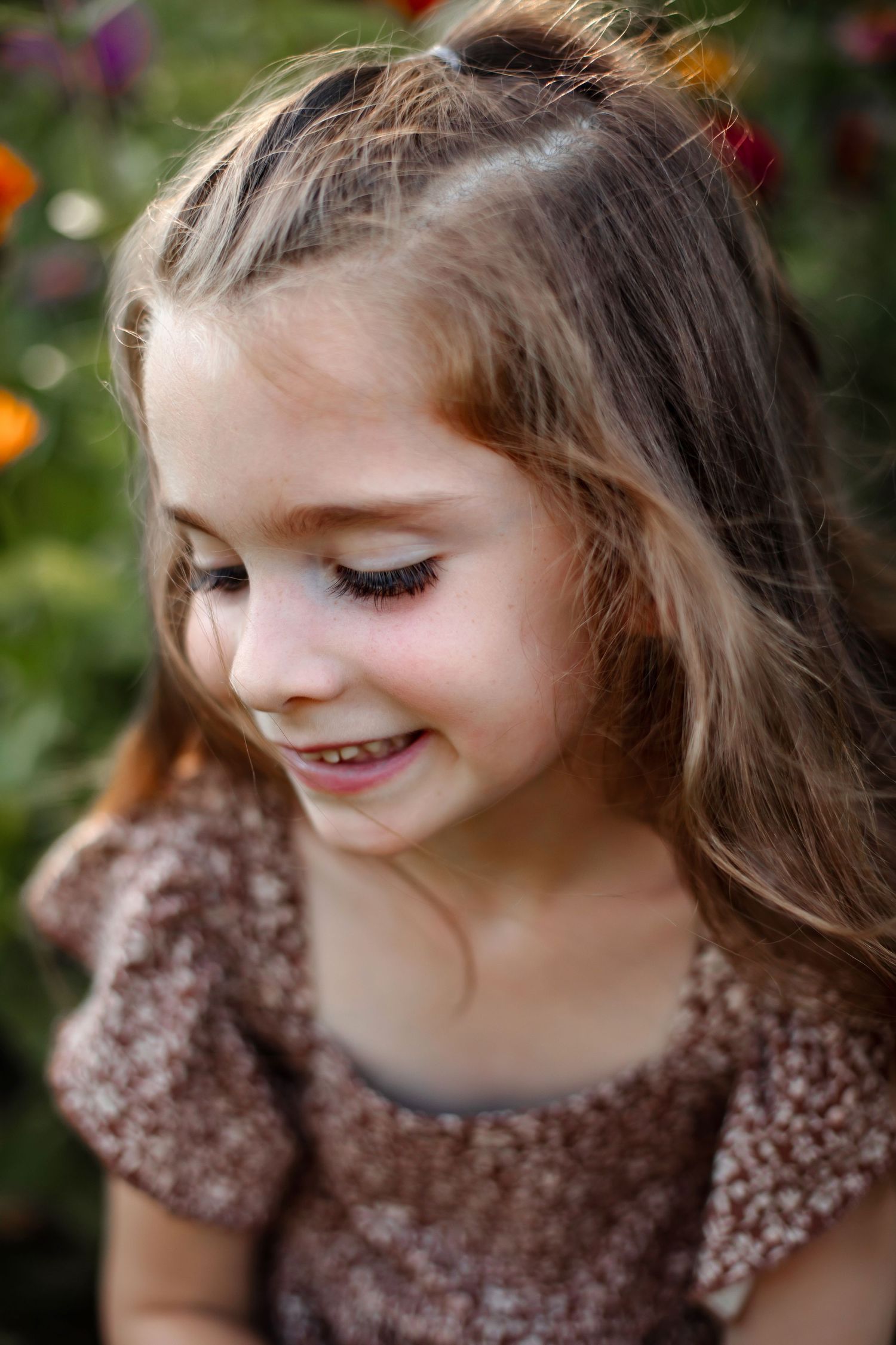 A young child wearing a brown sequined dress smiles while looking down amidst colorful flowers in a garden.