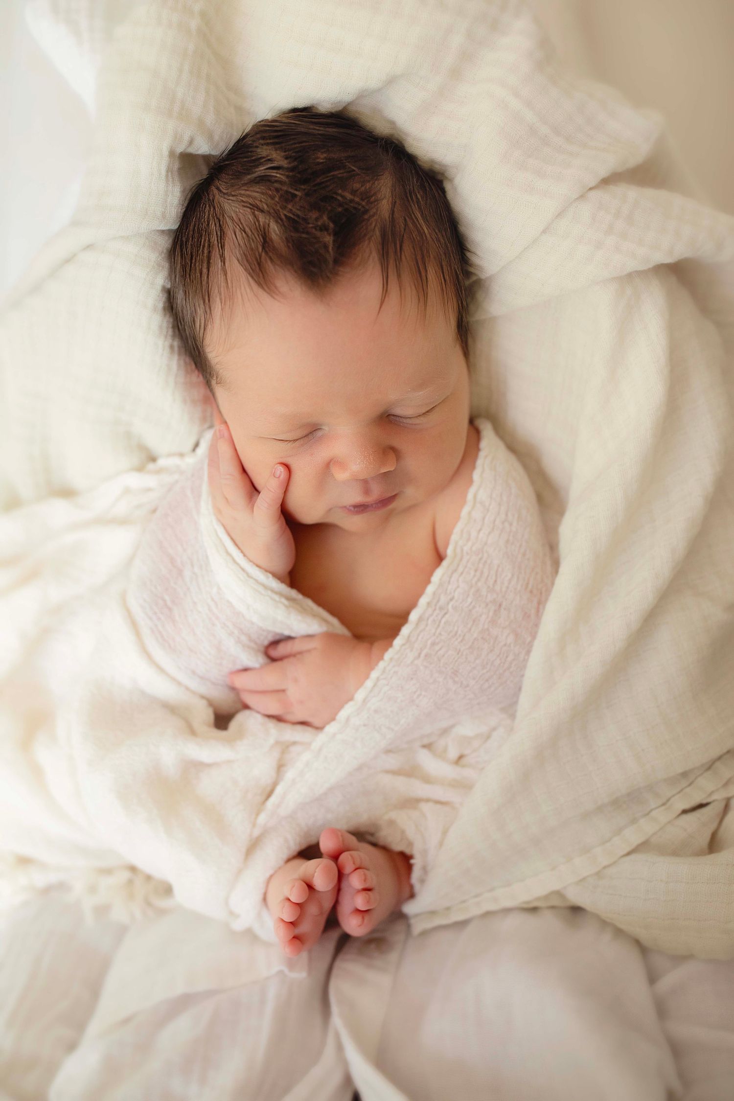 Peaceful newborn baby sleeping wrapped in soft white muslin blanket with tiny hands tucked under cheek.
