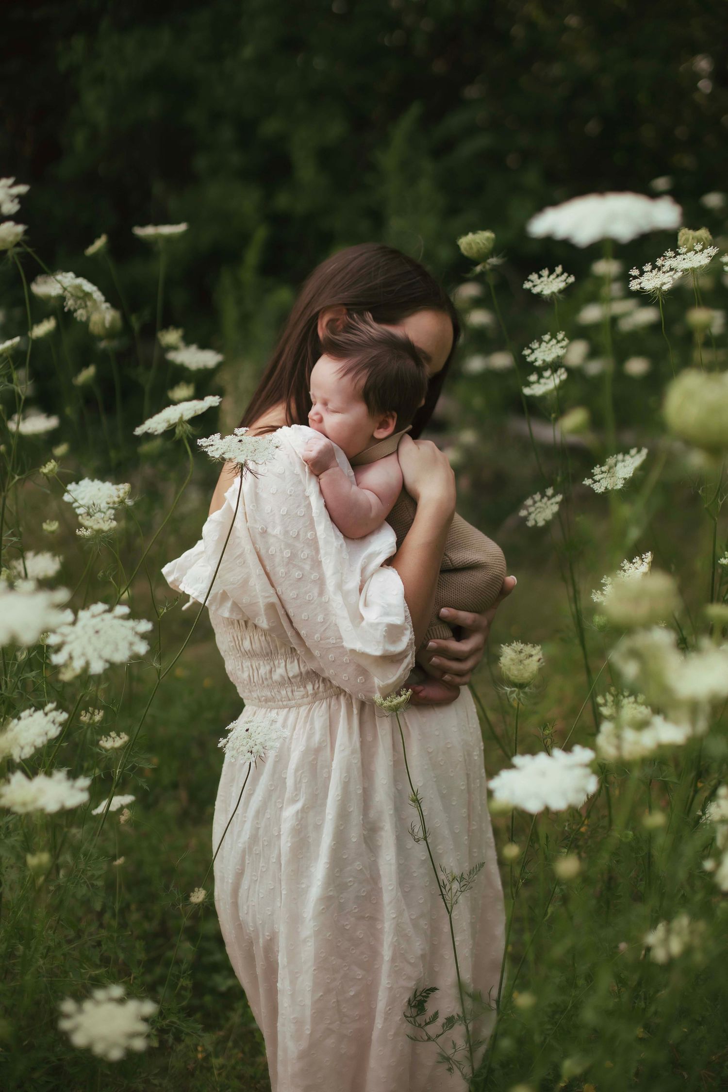 Young mother cradling newborn baby in white wildflower meadow, wearing elegant cream dress outdoors.