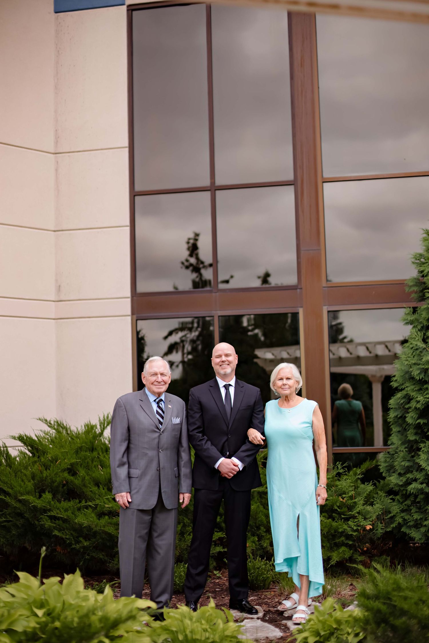 Three people in formal attire stand together outside a building with large windows and landscaping.