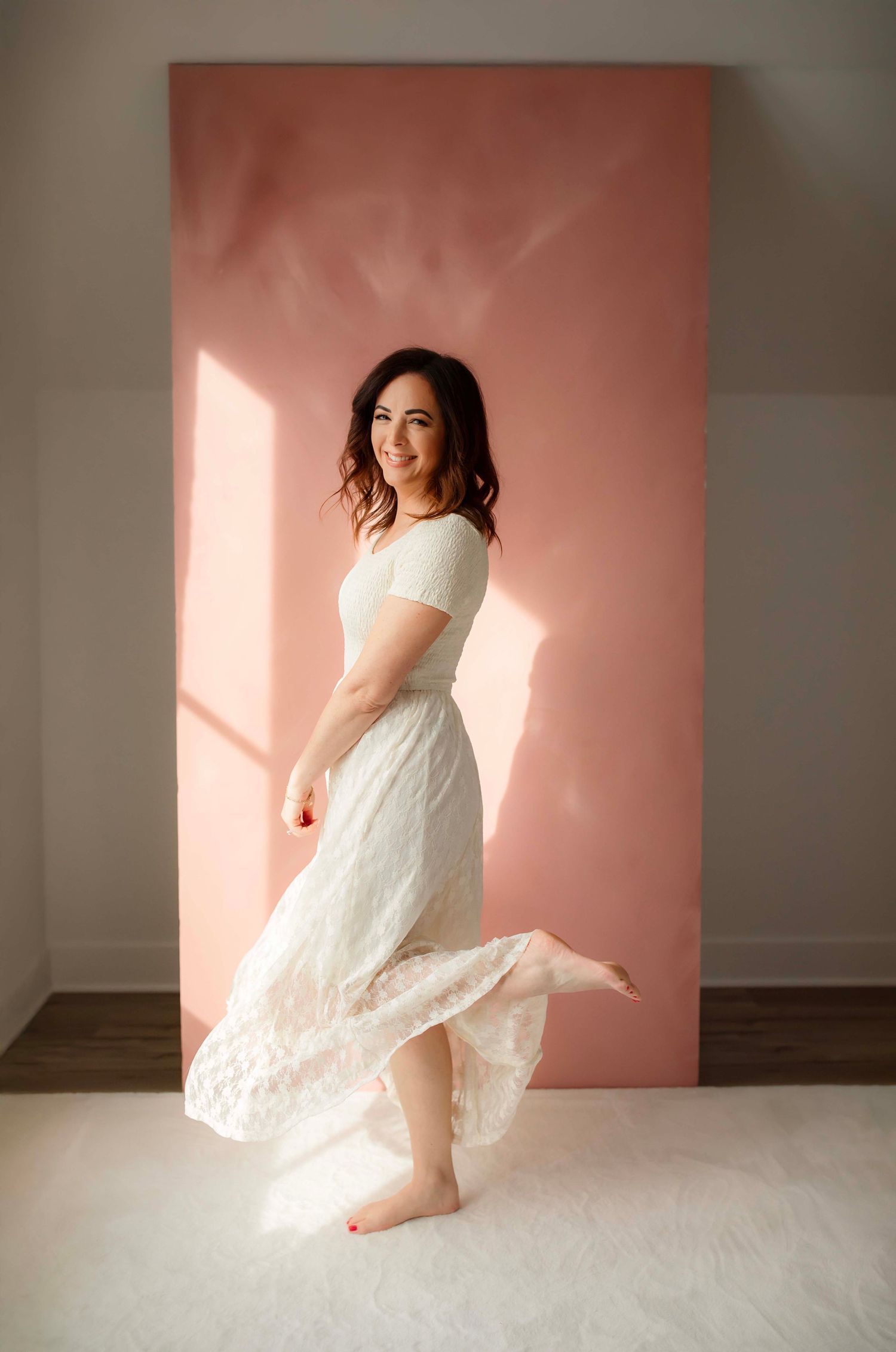 A figure in a flowing white lace dress twirls gracefully against a pink-tinted wall in a minimalist studio setting.