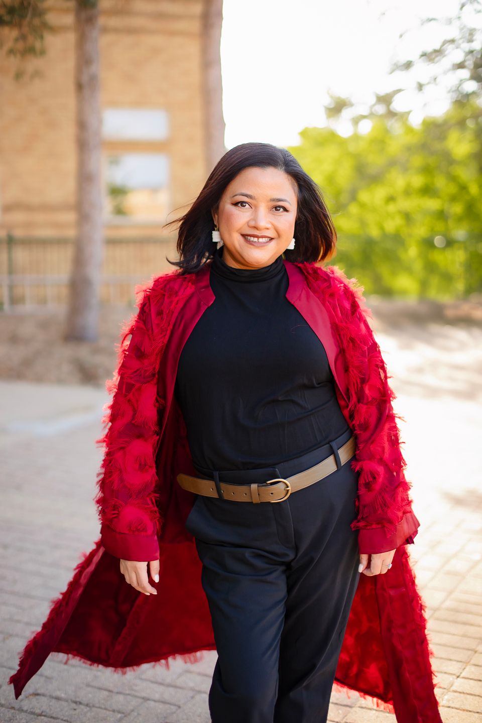 Someone wearing a stylish red faux fur coat over a black turtleneck outfit poses outdoors with a bright smile.