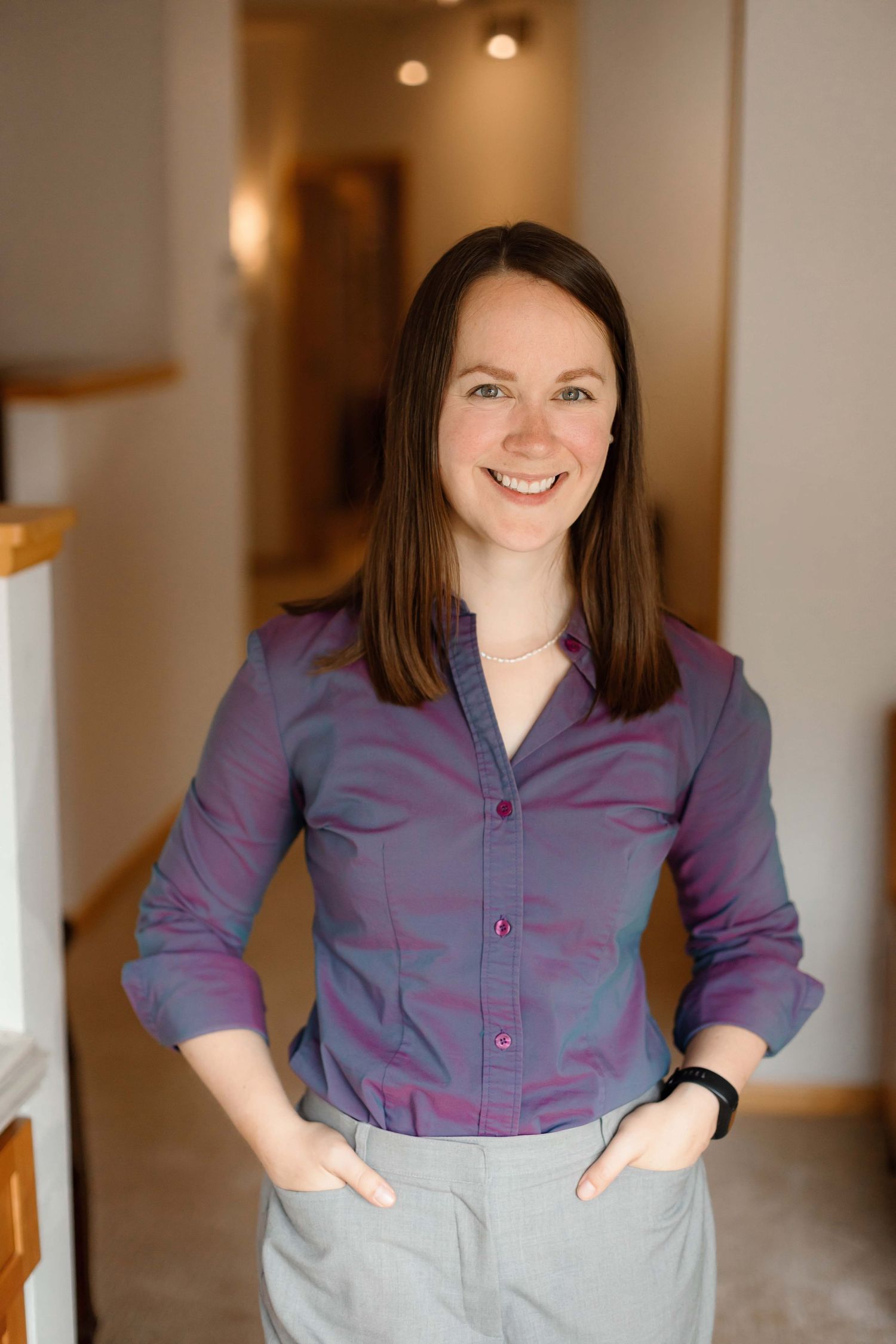 Professional woman in purple button-down shirt and gray pants stands in office hallway.