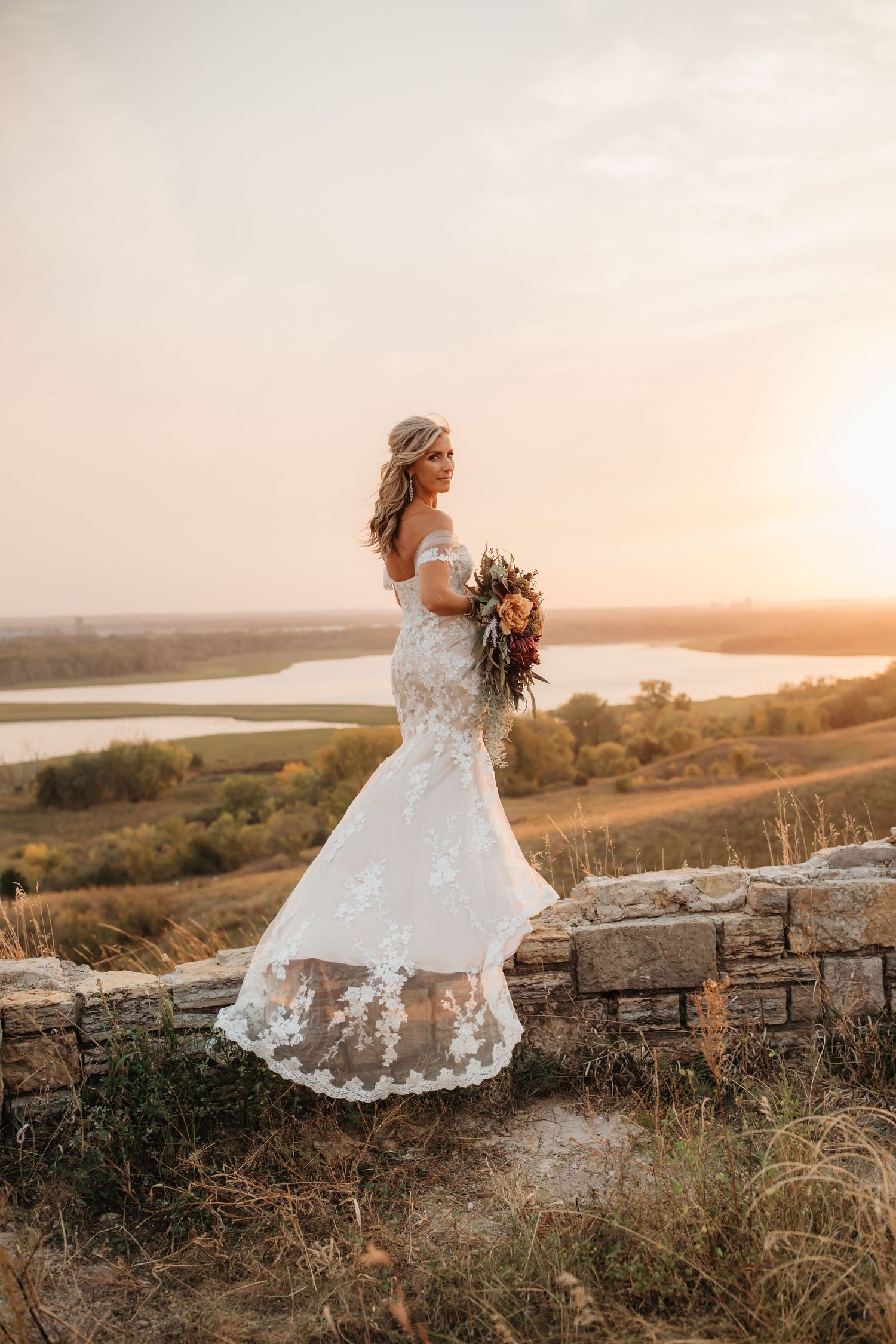 Bride in lace dress poses with bouquet on stone wall during sunset.