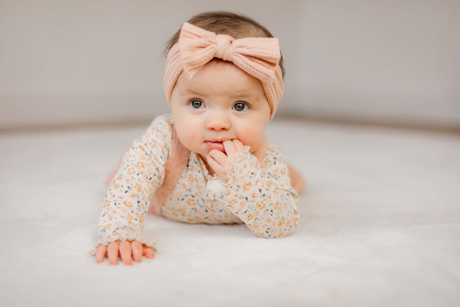 Baby in floral print outfit with pink bow headband lying on light carpet.